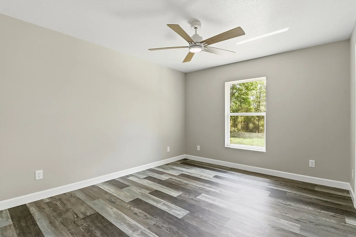 Empty room with beige walls, wood laminate flooring, a window showing green trees outside, white ceiling fan, and white baseboards.