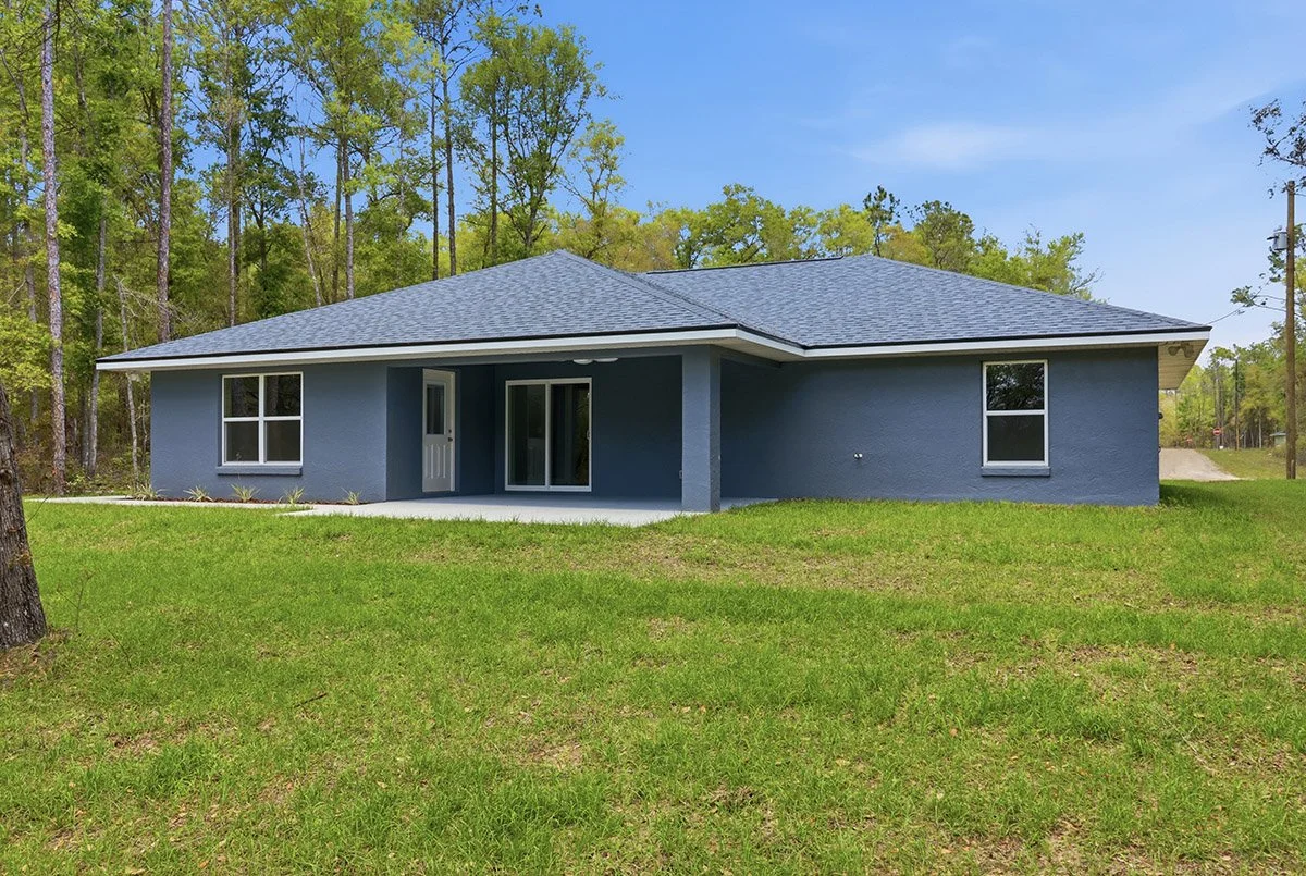 A single-story blue house with a gray roof, large windows, and a small covered porch, surrounded by green grass and trees.