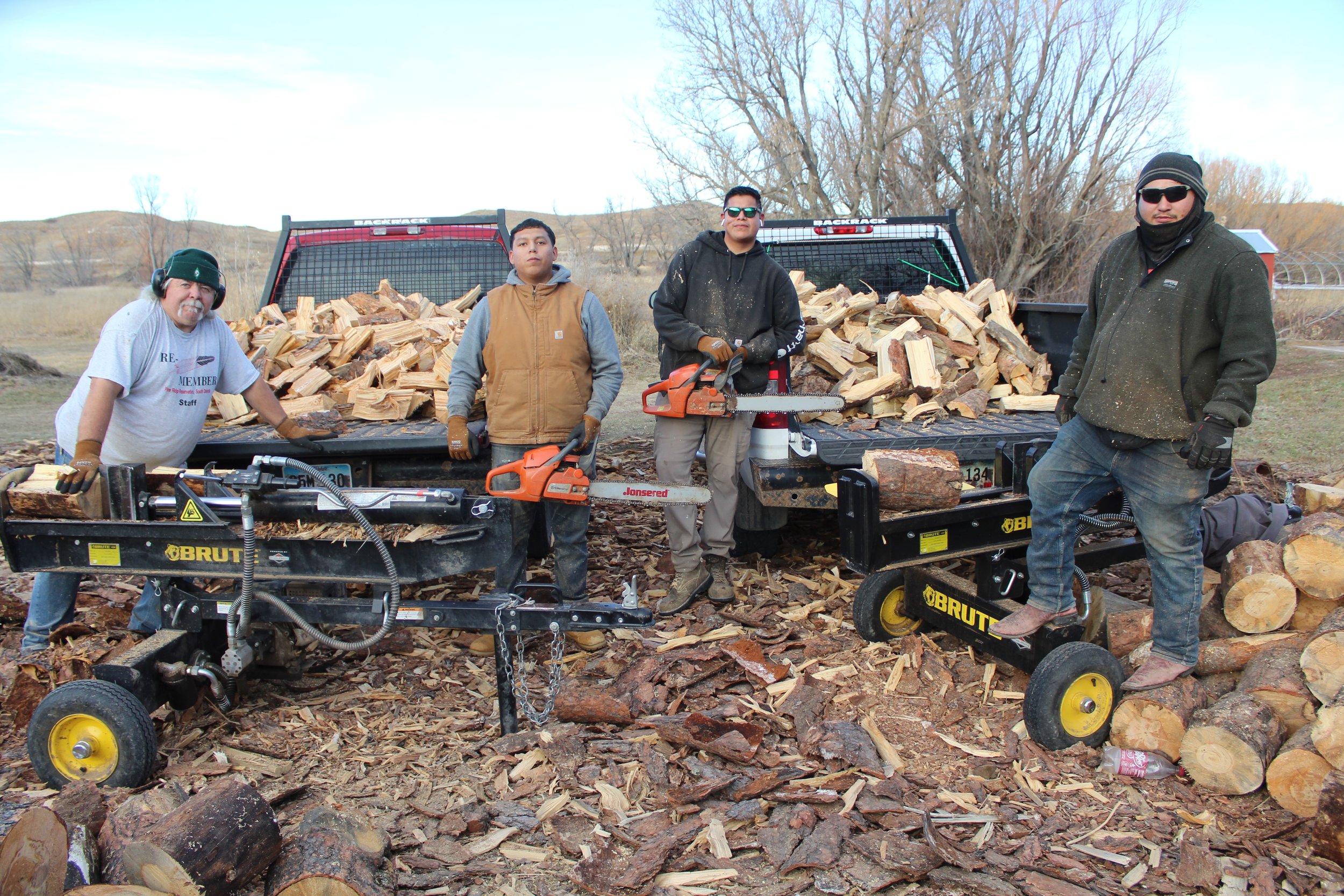 Four men stand at the Re-Member woodpile with splitters and chainsaws