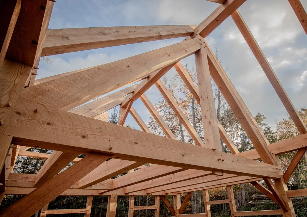 Timber frame construction under blue sky and clouds, showing wooden beams and trusses, surrounded by trees.