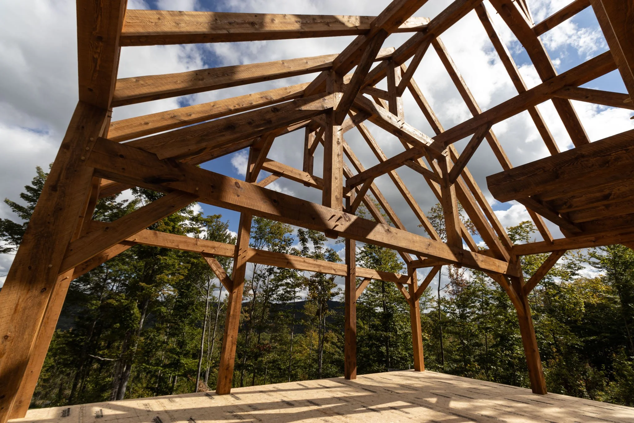 Wooden timber frame of a structure under construction outdoors, surrounded by trees and a cloudy sky.