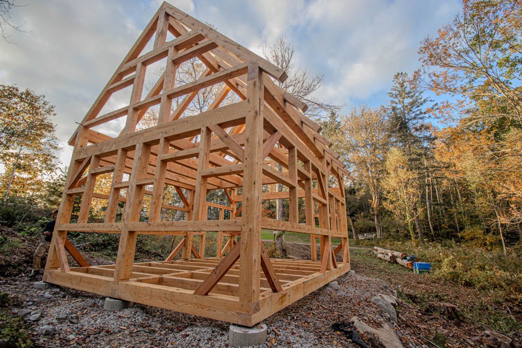 Wooden timber frame structure under construction in a forested area with autumn foliage.