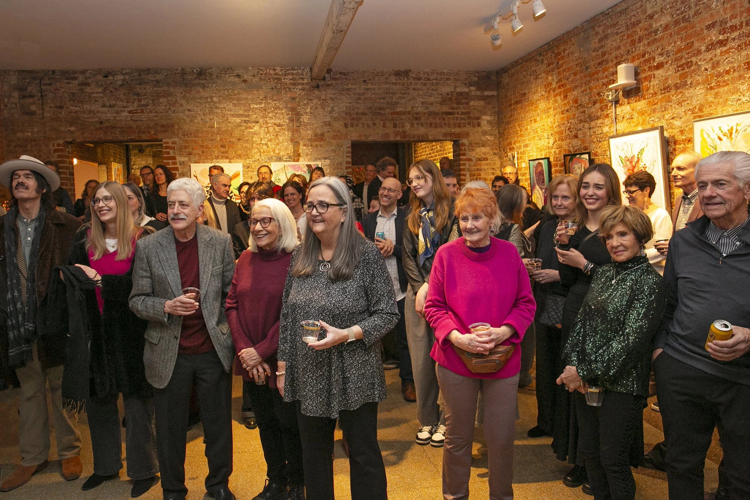 Group of people gathered at an art gallery event, smiling and holding drinks, with colorful artwork displayed on brick walls in the background.