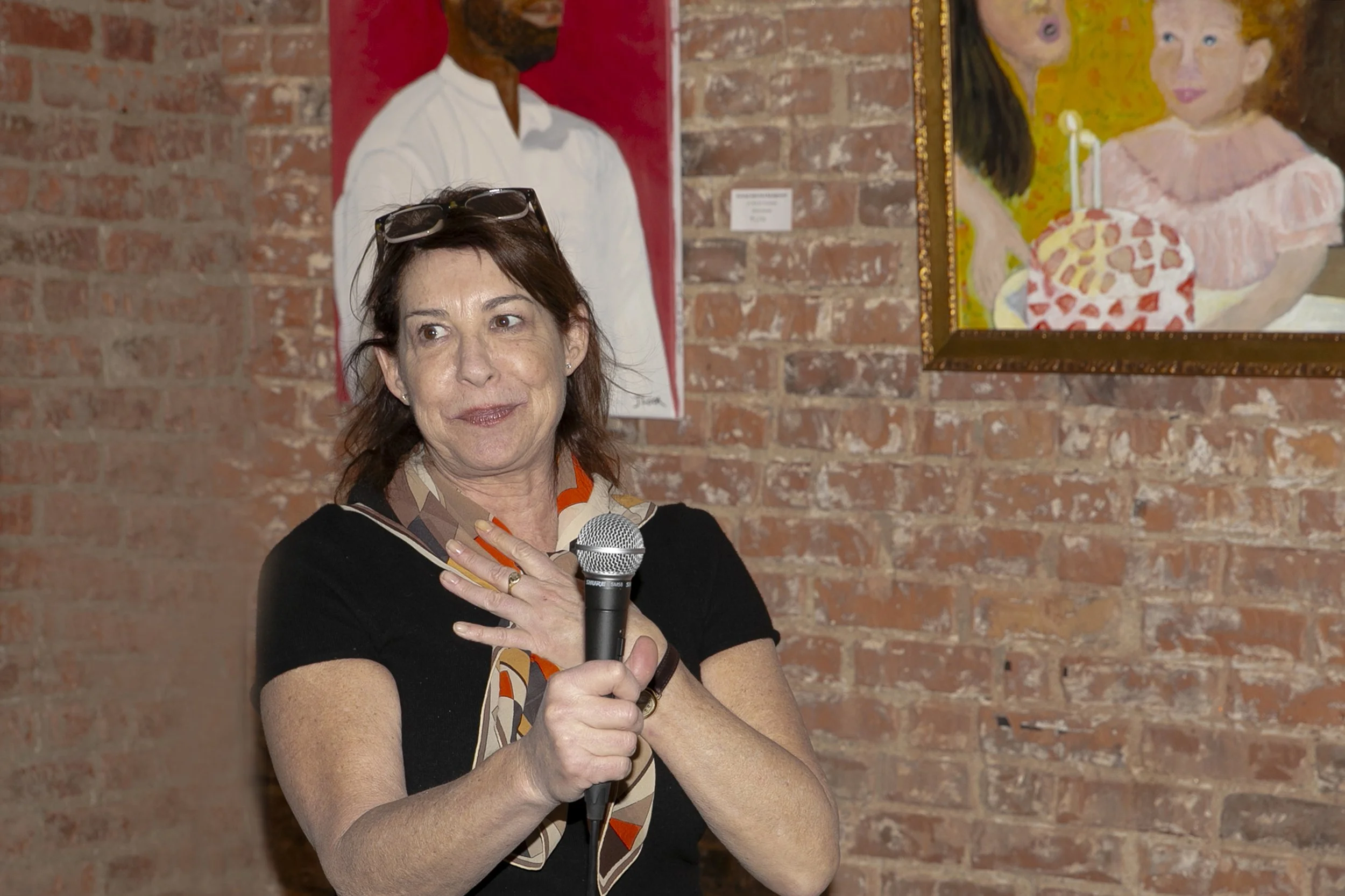 A woman with glasses on her head holding a microphone near her mouth, standing in front of a brick wall with colorful paintings hanging on it.
