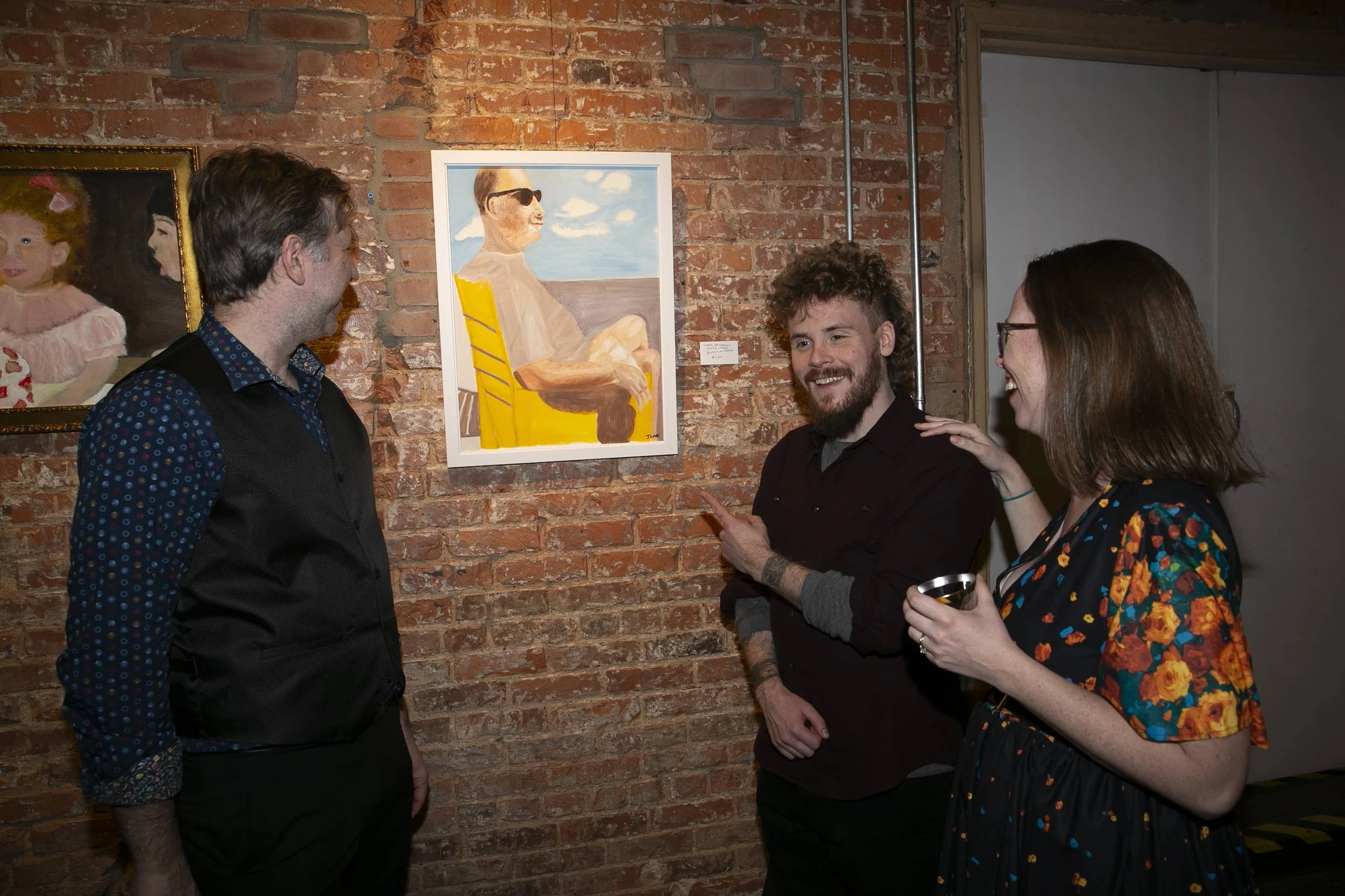 Three people at an art gallery look at and discuss a painting of a man sitting in a yellow chair on a brick wall background.