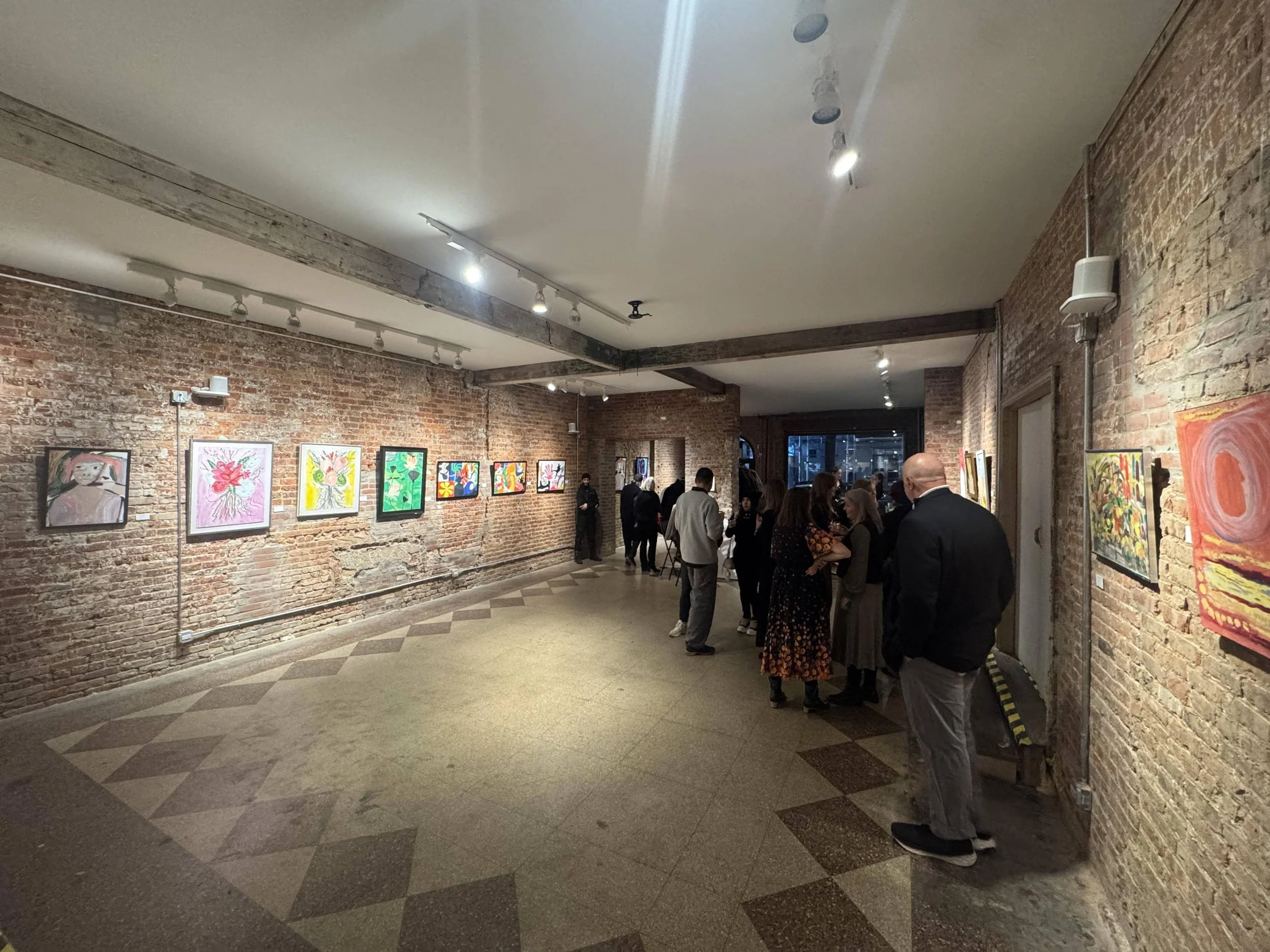 People viewing colorful artwork on brick walls inside an art gallery with warm lighting.