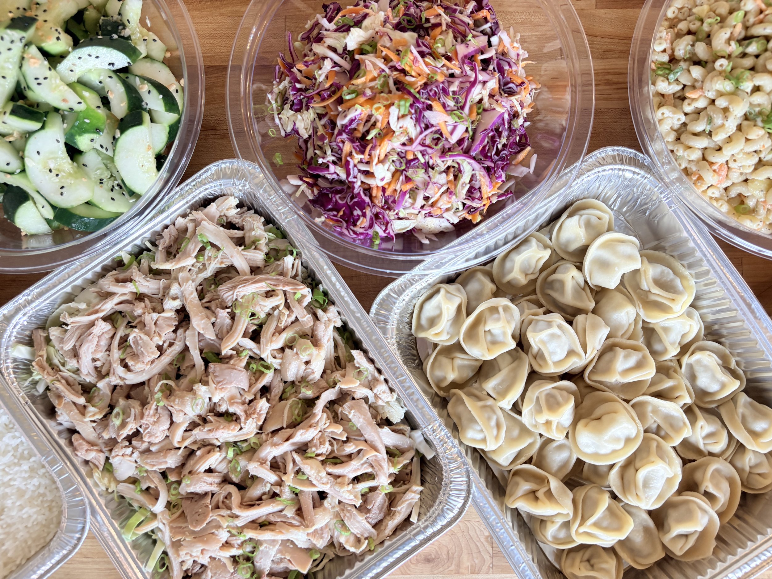 catering display consisting of cucumber salad, pulled chicken, white rice, rainbow miso slaw, pork dumplings, and hawaiian mac salad