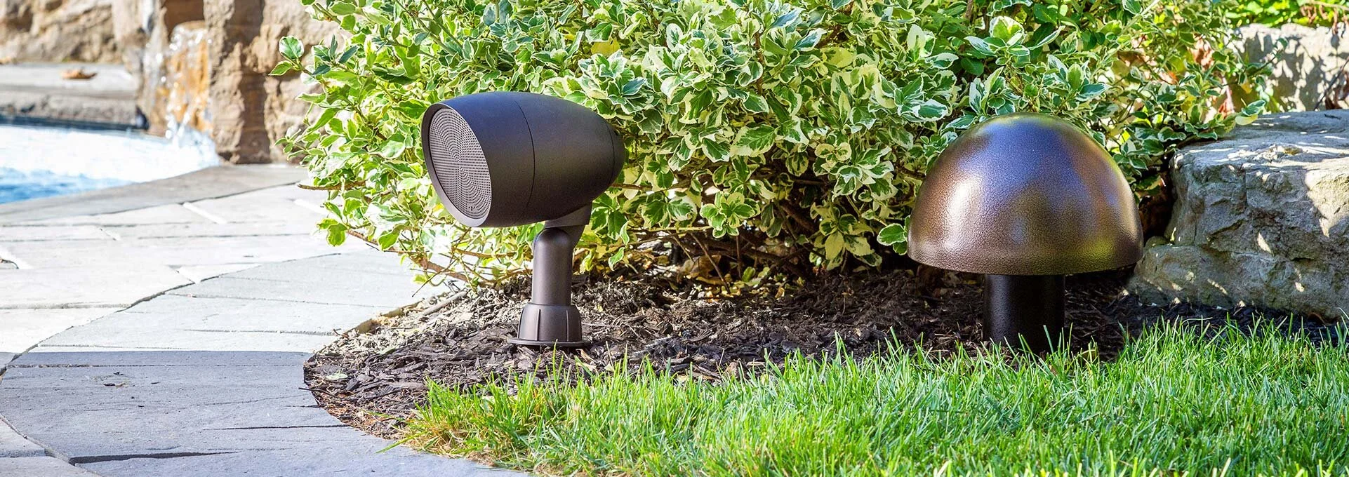 Outdoor landscape with a pathway, a bush, and outdoor lighting fixtures, including a black spotlight and a decorative mushroom-shaped light, near a stone wall and grass.