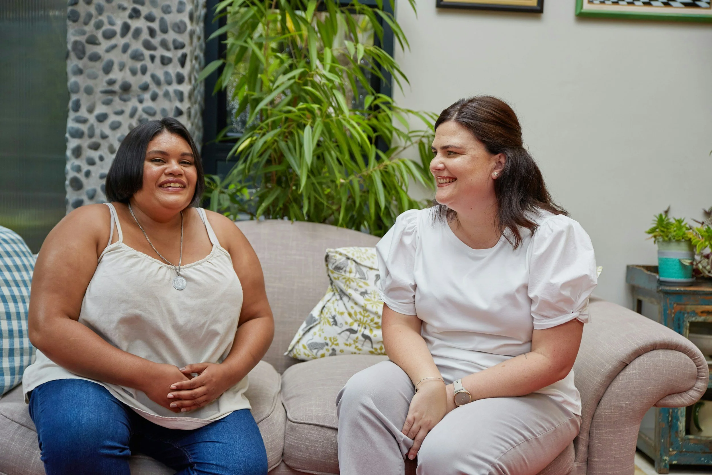 Two female presenting adults sitting on a couch with a green plant in the background. They both have smiles on their faces and appear happy.