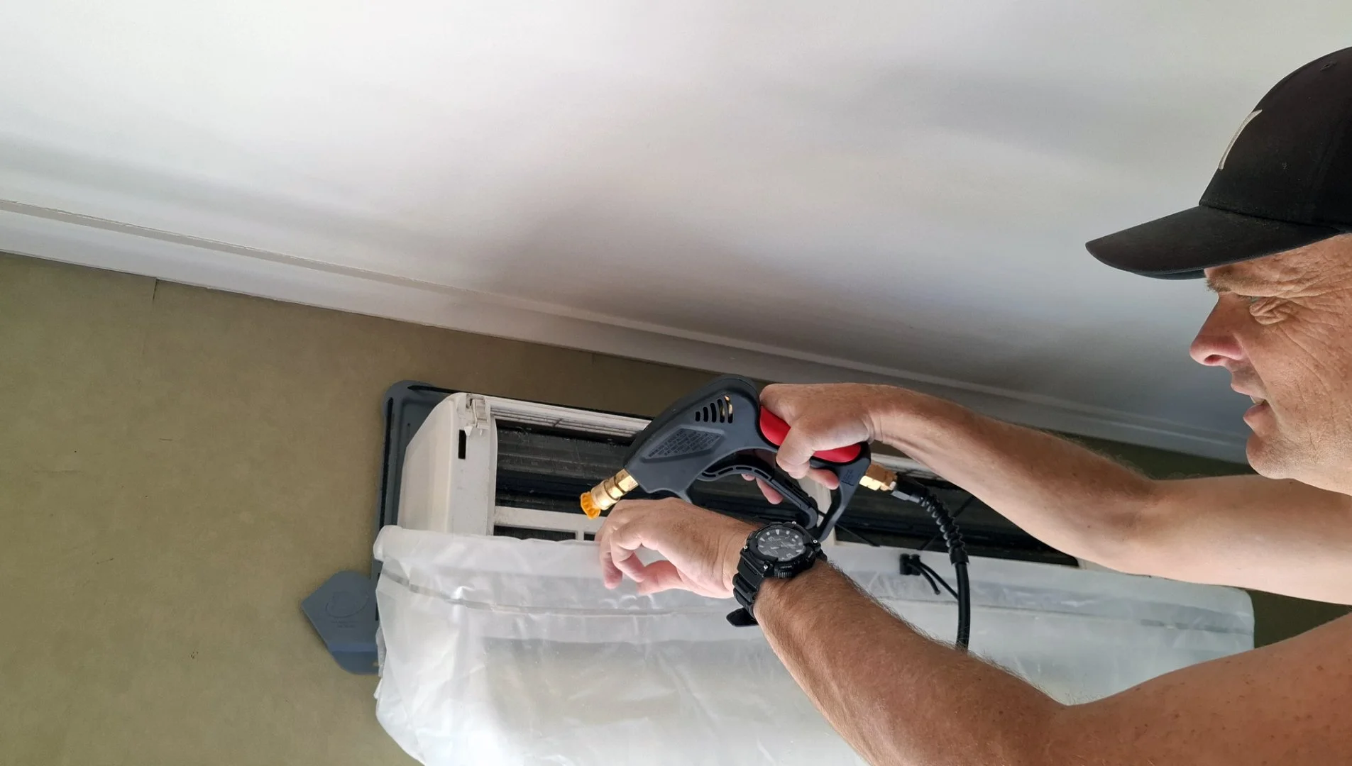 A man in a black cap is cleaning an air conditioning unit with a handheld pressure washer.