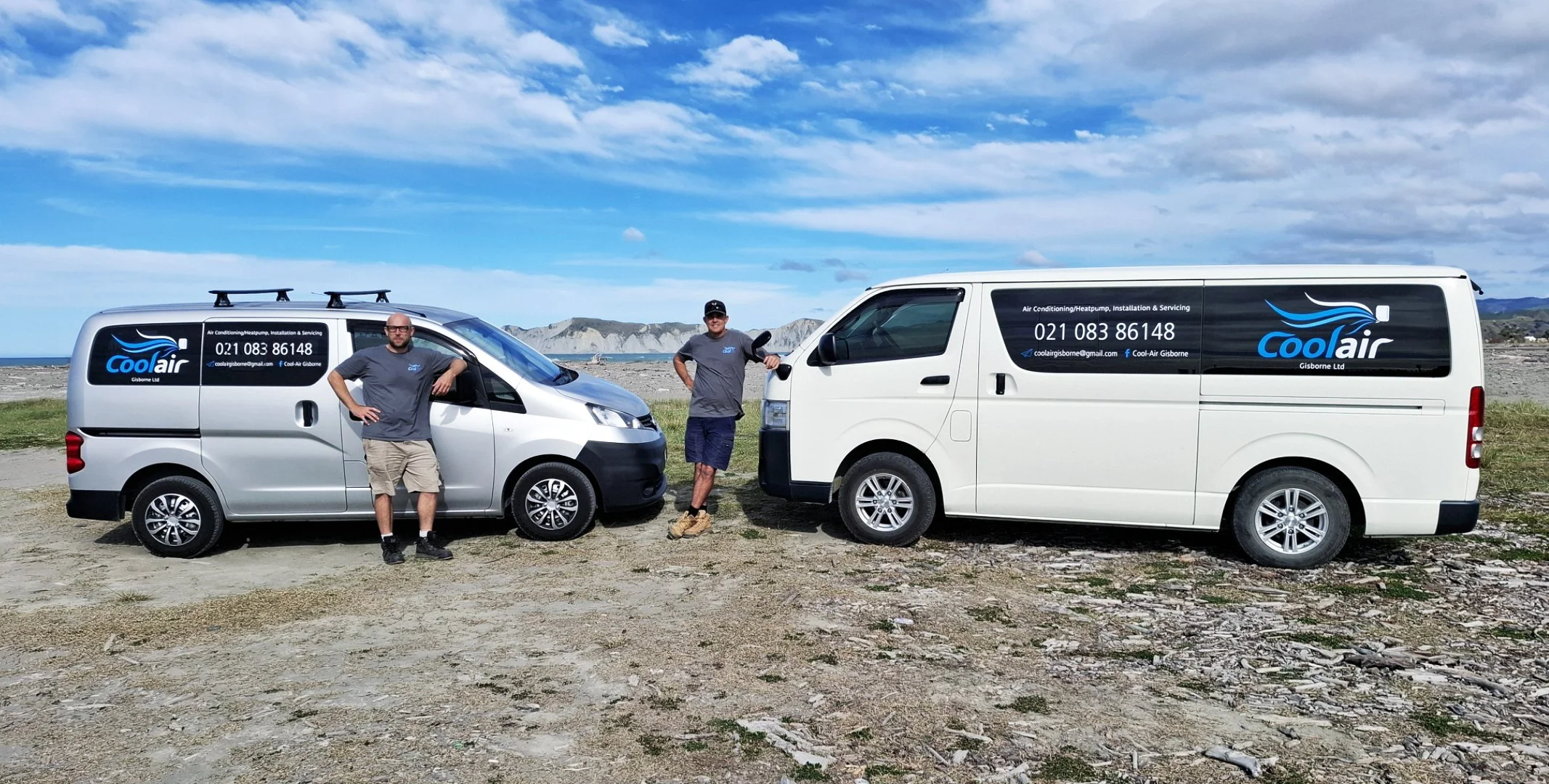 Two men standing next to company vehicles on a rocky beach with mountains in the background. Both vehicles display a logo and contact information for Coolair, an air conditioning and heat pump service company.