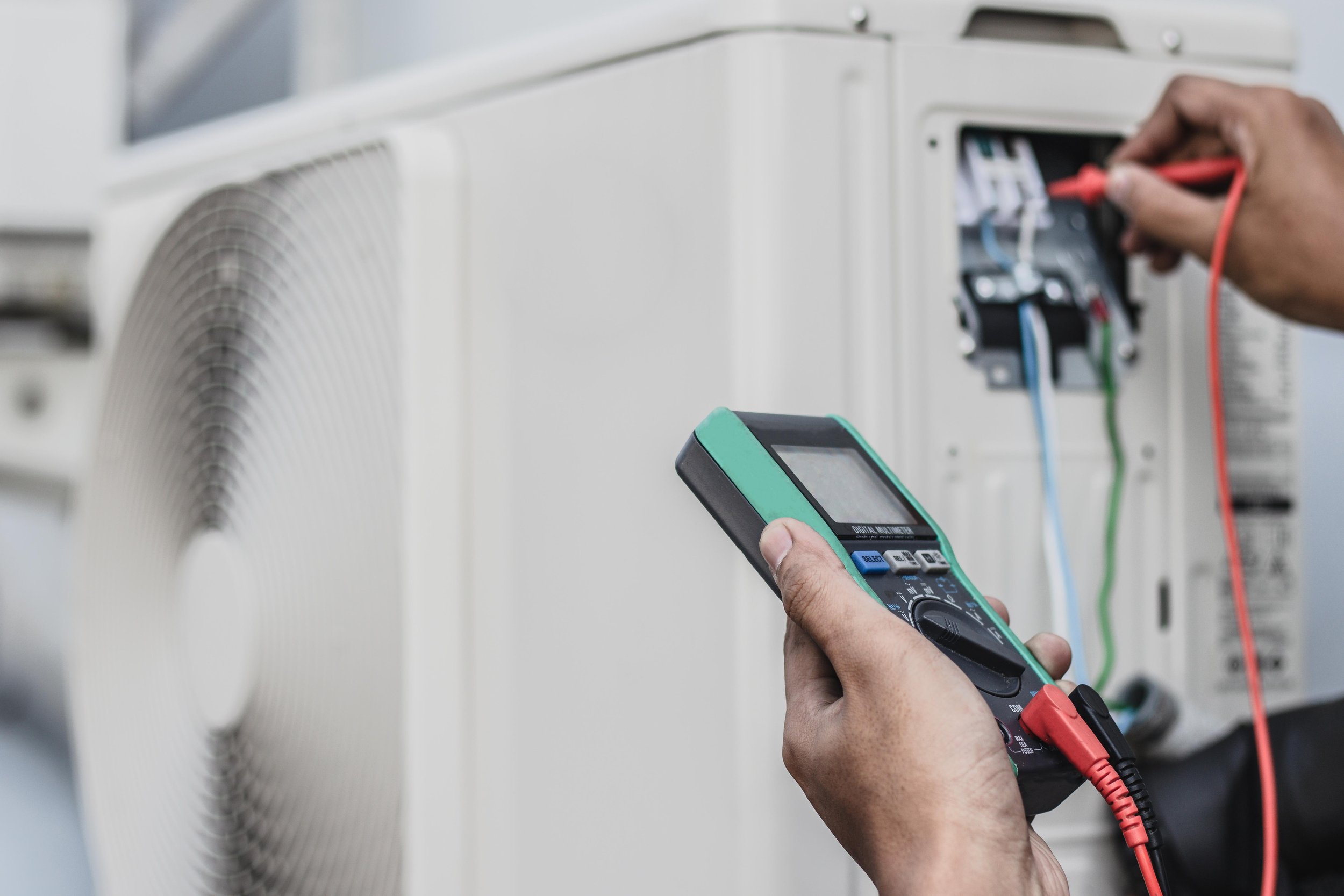 A person holding a digital multimeter testing wires connected to an air conditioning unit.