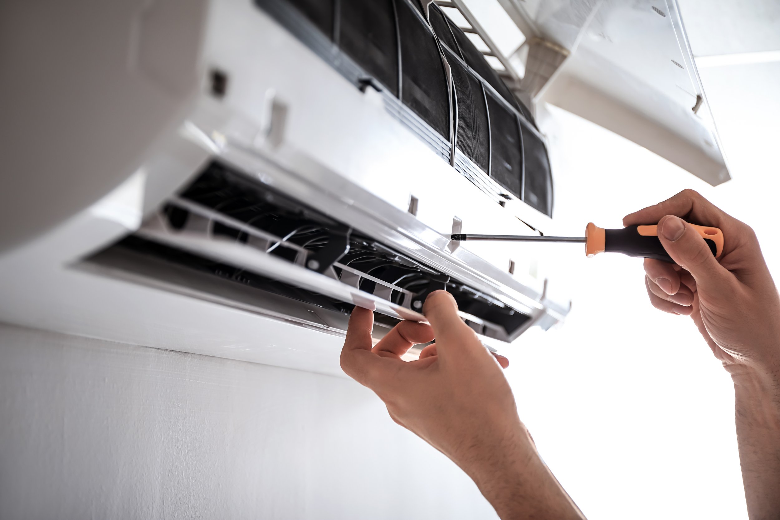 Person using a screwdriver to remove or install a part inside an air conditioning unit or ventilation system on a white wall.