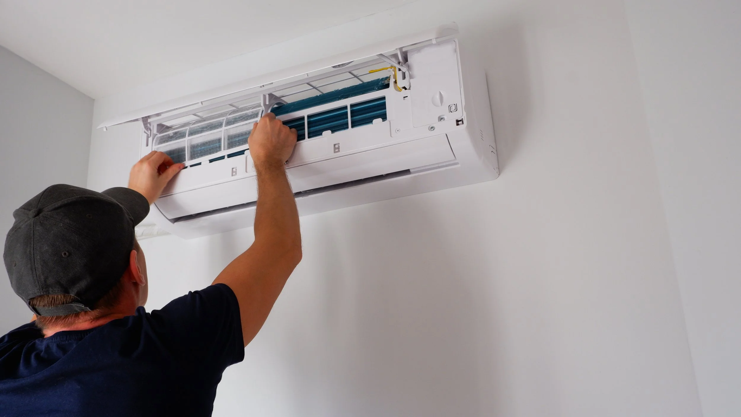 A man in a dark shirt and baseball cap installing or repairing a wall-mounted air conditioner, working on the interior unit of the system.