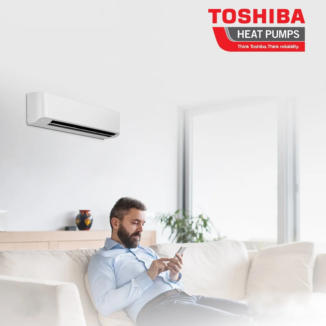 A man sitting on a white sofa in a bright living room, looking at his phone. An air conditioning unit is mounted on the wall above him. The room has a large window with sunlight coming in, and there is a potted plant and a vase in the background. The