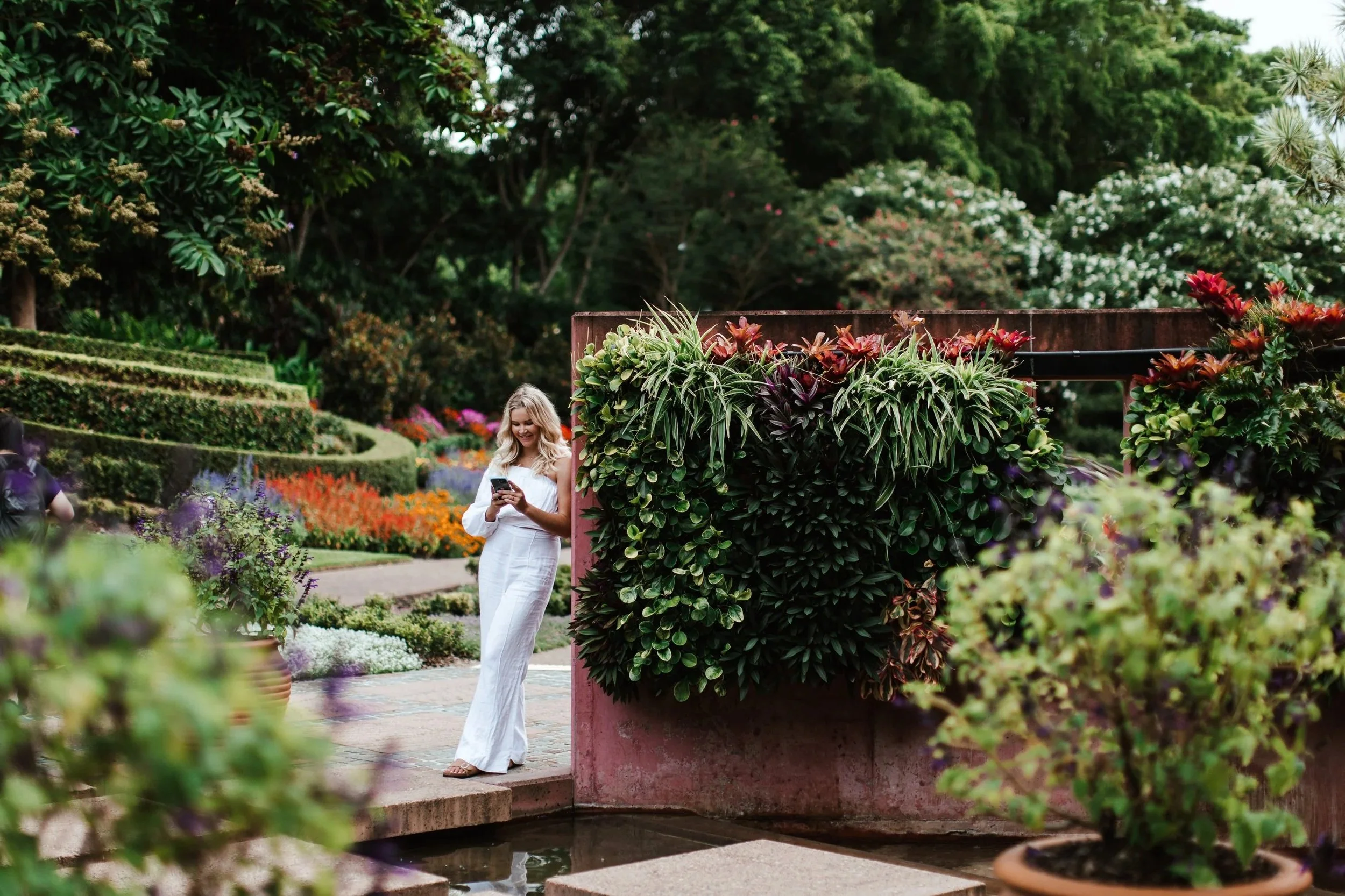A woman in white jumpsuit standing in a lush, colorful garden, looking at her phone near a small water feature.