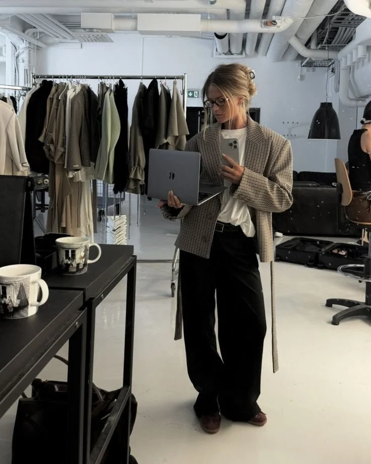 A woman taking a mirror selfie in a fashion studio or boutique, holding a laptop and smartphone, with clothing racks of neutral-colored garments behind her, and coffee mugs on a black table.