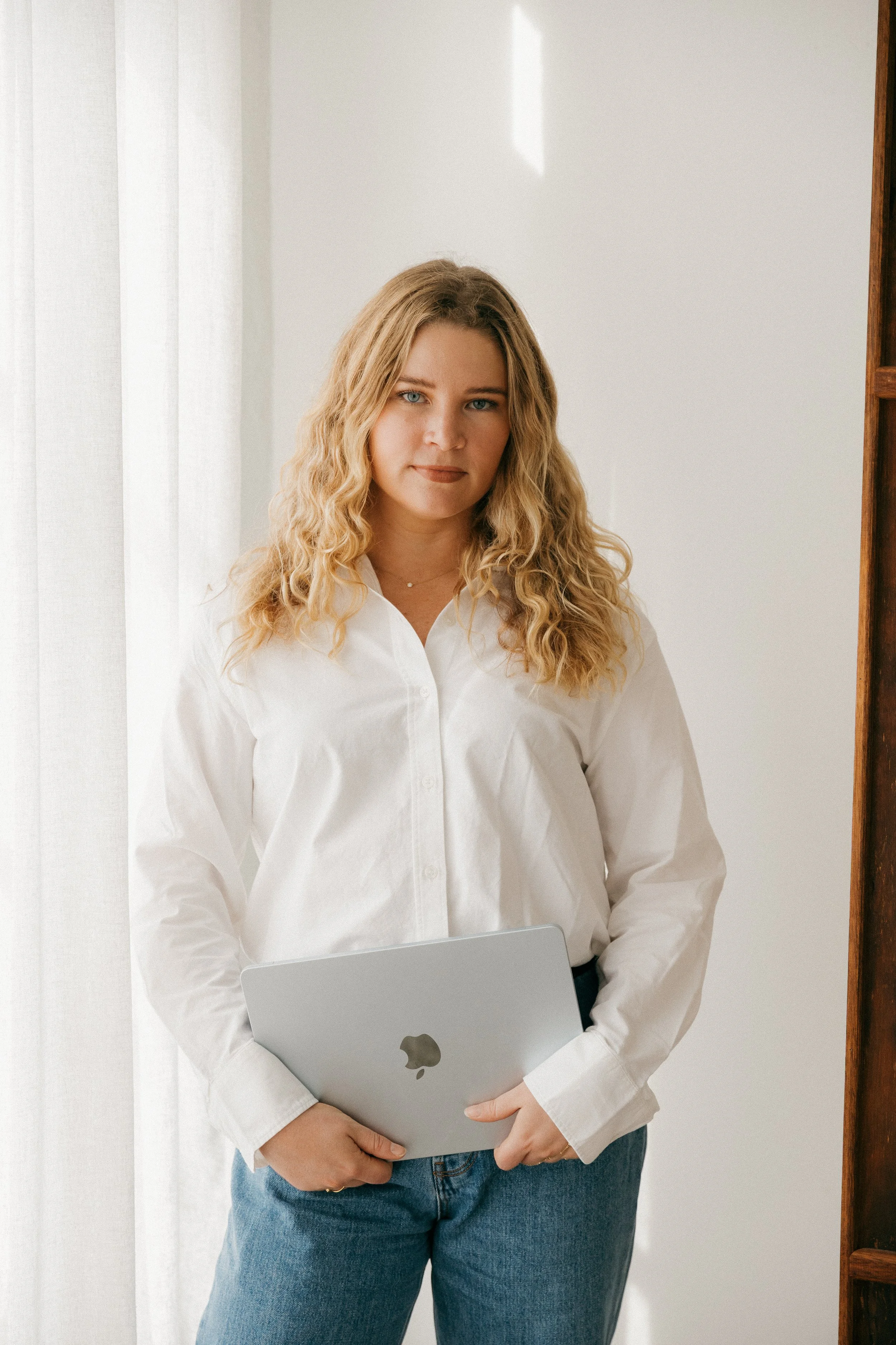 A woman with curly blonde hair wearing a white shirt holding a silver laptop, standing in front of a white wall with a wooden bookshelf to the right.