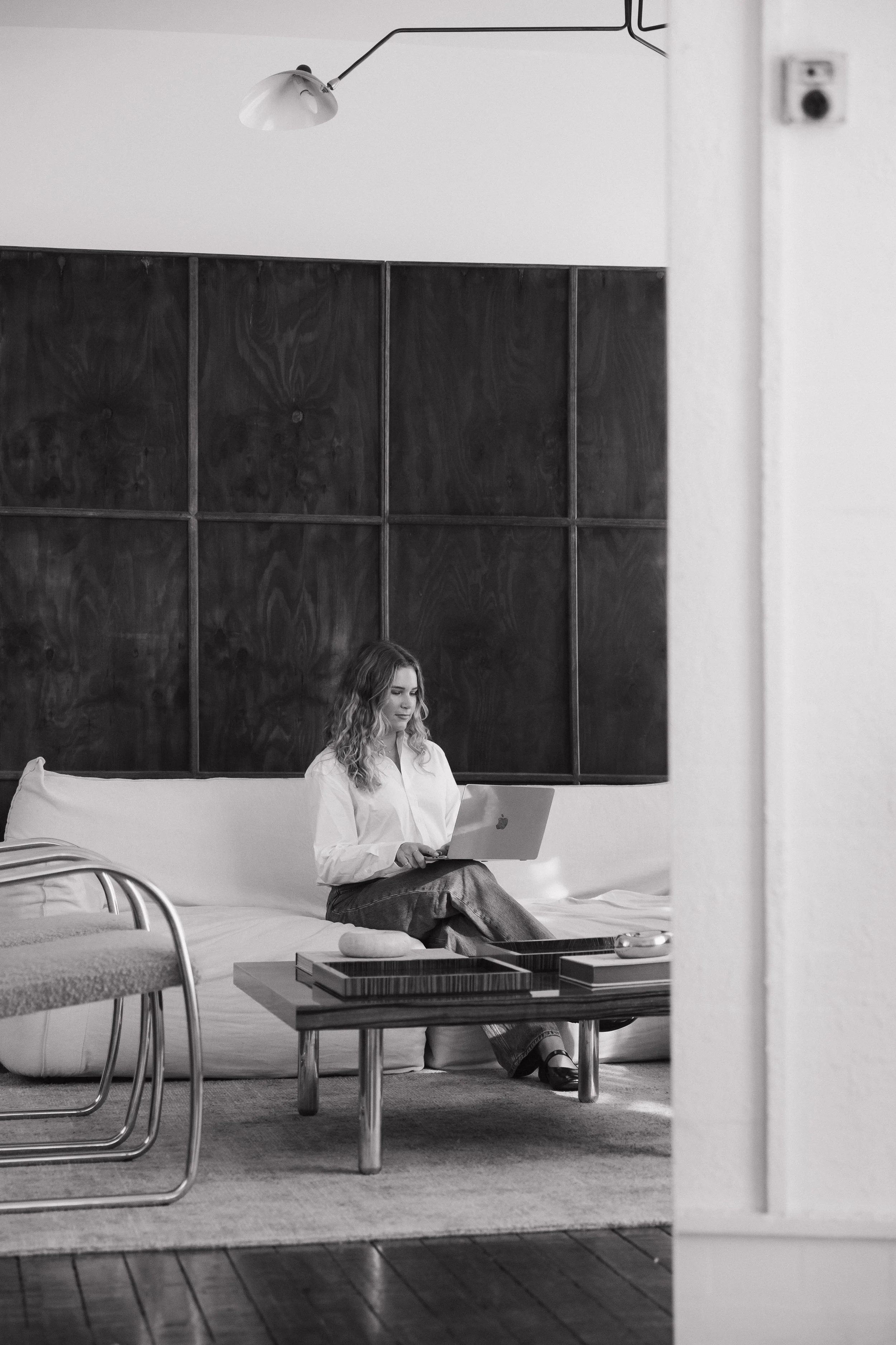 A woman with curly hair sitting on a bed, working on a laptop in a modern, minimalist room with a dark wood-paneled wall and a white wall.