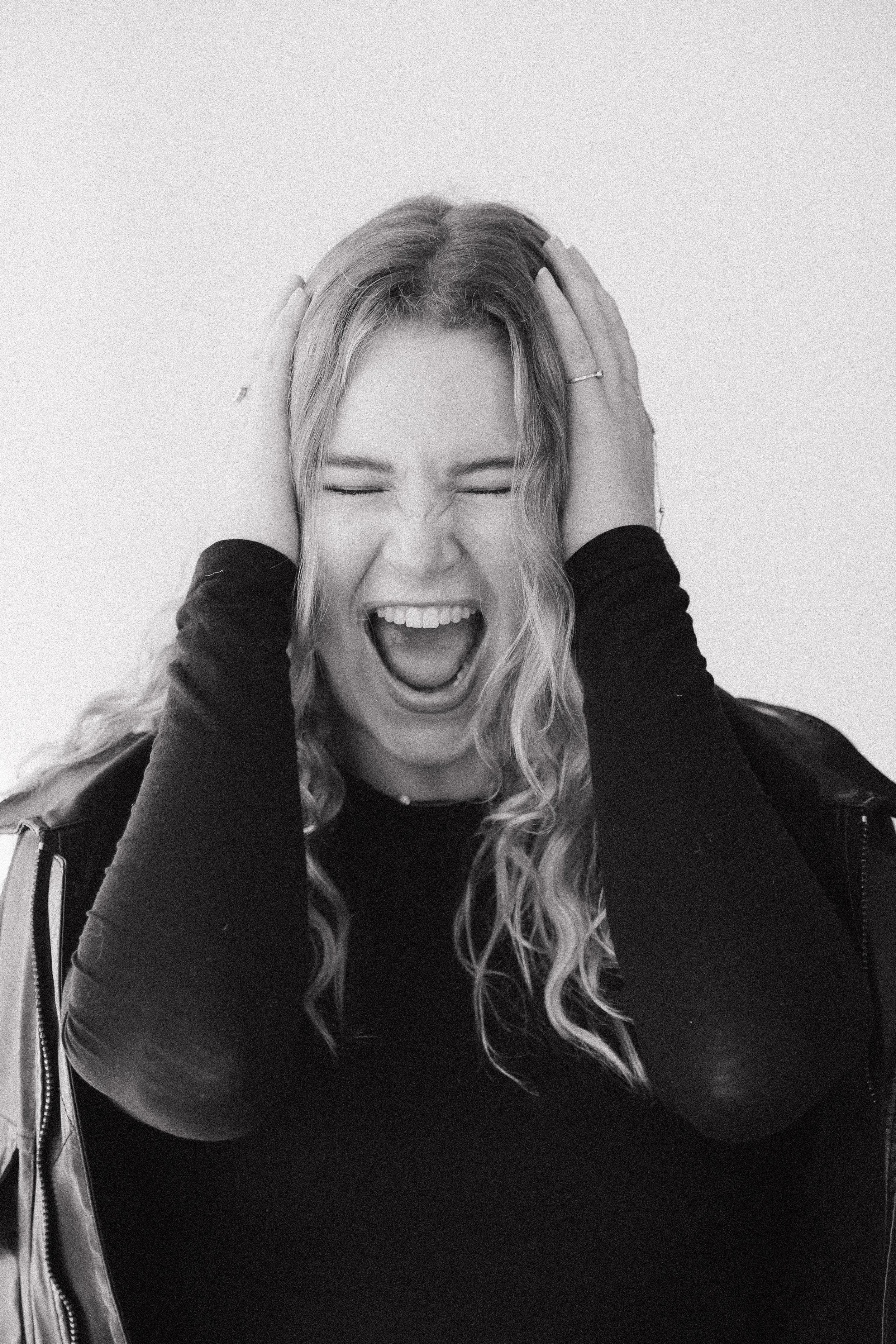 A woman with long wavy hair, wearing a black long-sleeve shirt, is holding her head with both hands and appears to be screaming or shouting with her eyes closed and mouth open, against a plain background.