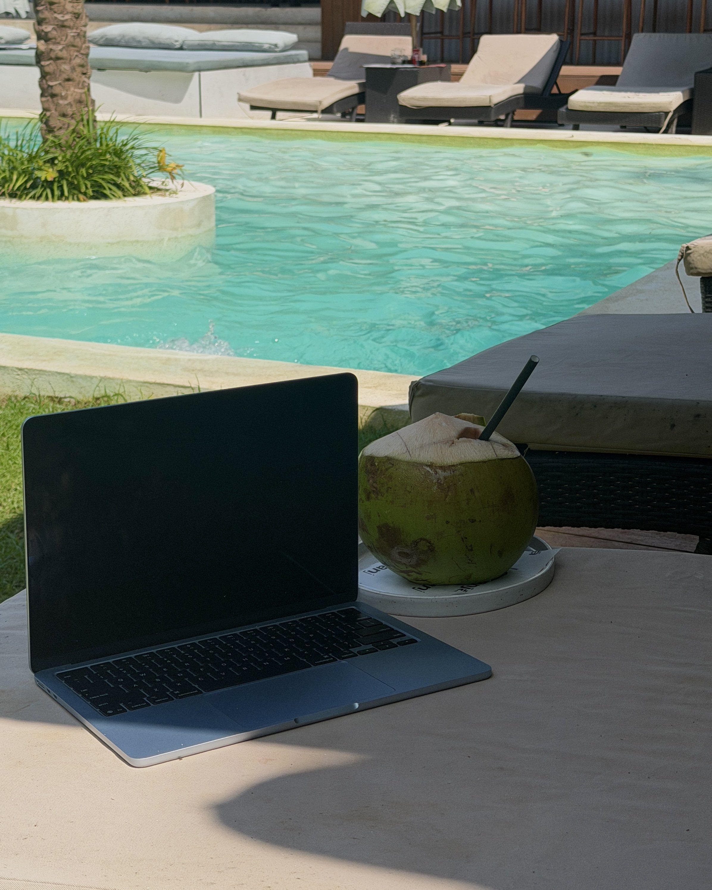 Laptop, coconut drink with straw on a tray, and poolside lounge chairs at a swimming pool  showing the lifestyle of a online freelancer digital nomad.