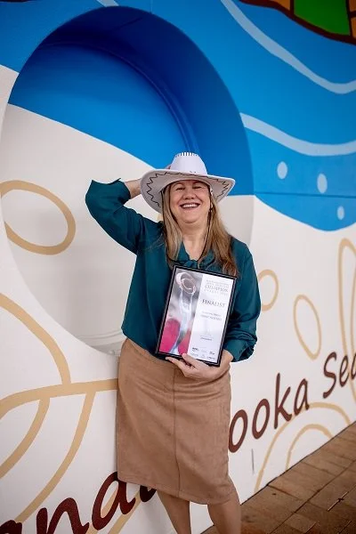 Woman smiling and holding a certificate, wearing a white cowboy hat and blue jacket, standing in front of a colorful sea-themed mural.