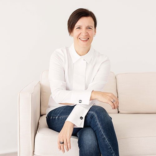 Smiling woman with short dark hair sitting on a white sofa against a light background, wearing a white shirt and blue jeans.