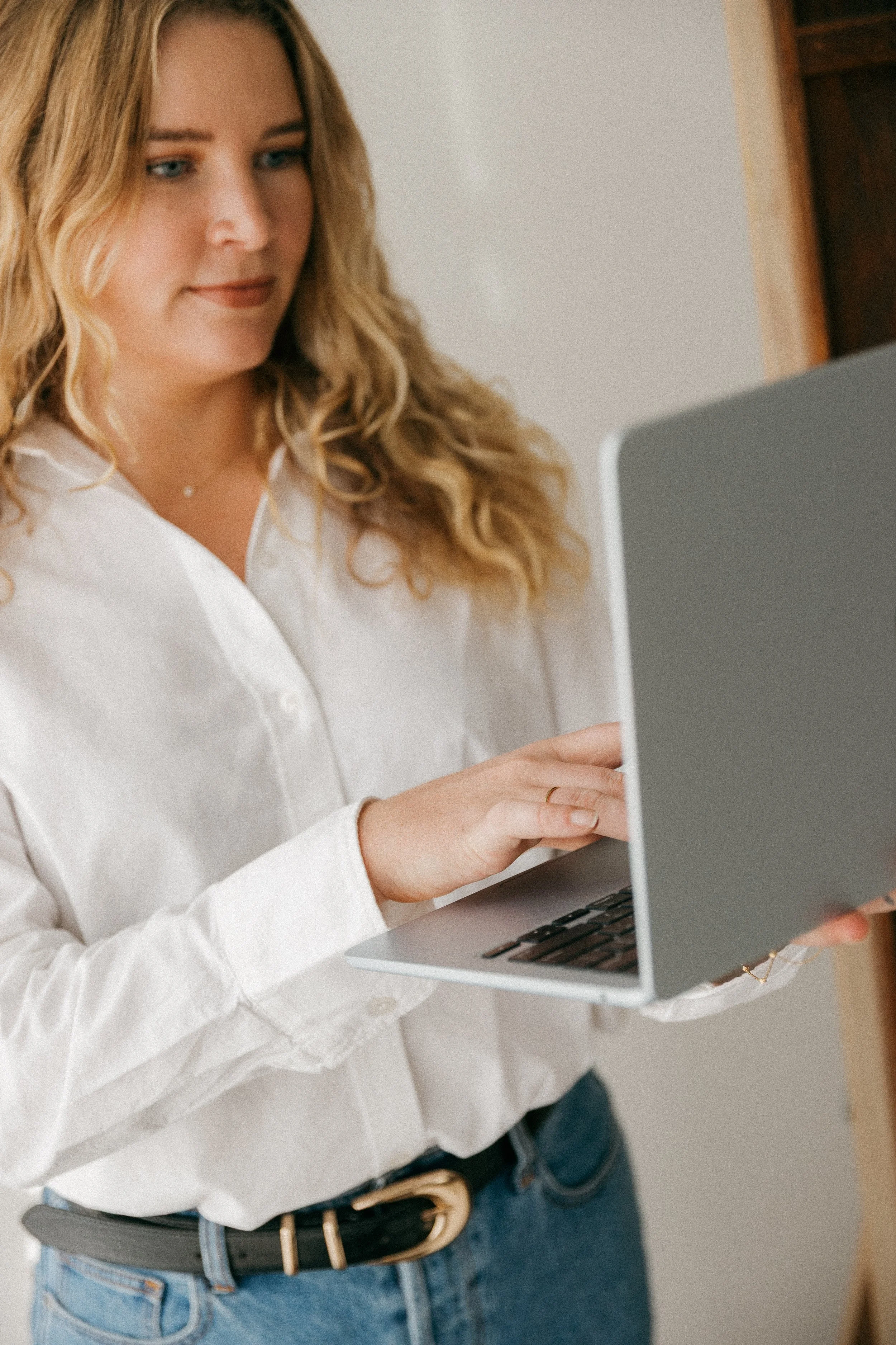 A woman with wavy blonde hair wearing a white blouse and blue jeans using a laptop.