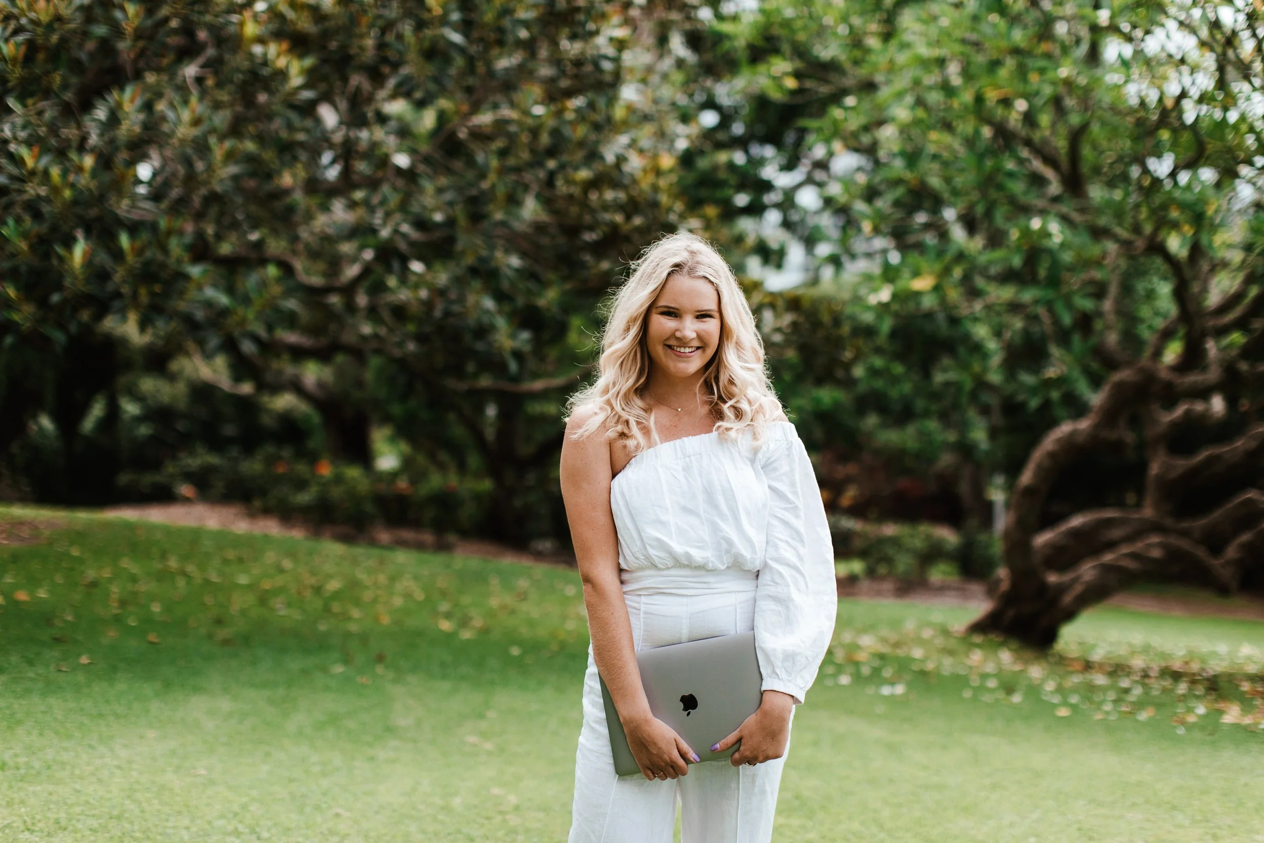 Hannah, founder of Jayne Meadia holding a closed silver laptop and smiling at the camera, with green trees and grass in the background.