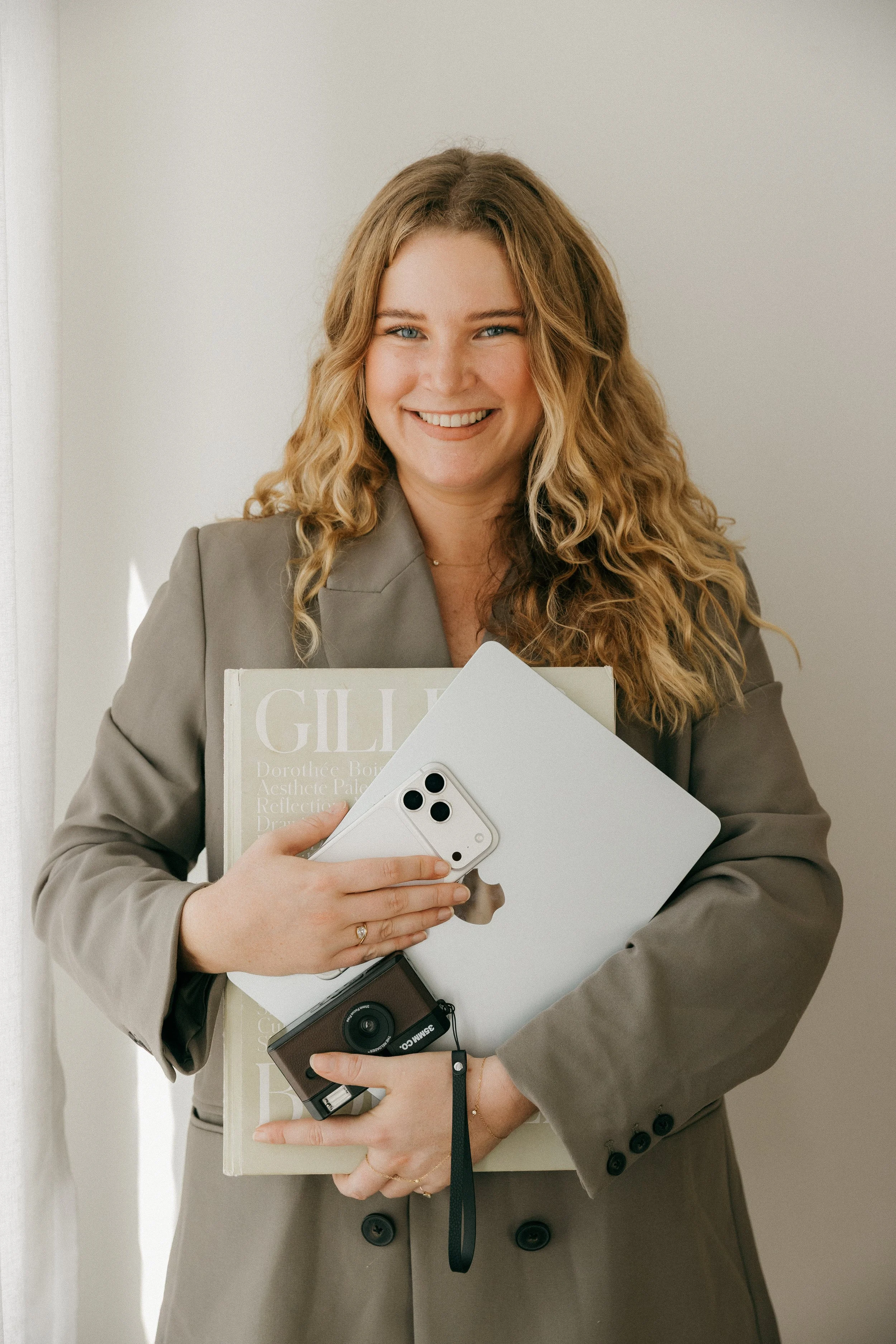 A woman with long curly blonde hair, wearing a gray blazer, is smiling and holding a tablet, a smartphone and a magazine.