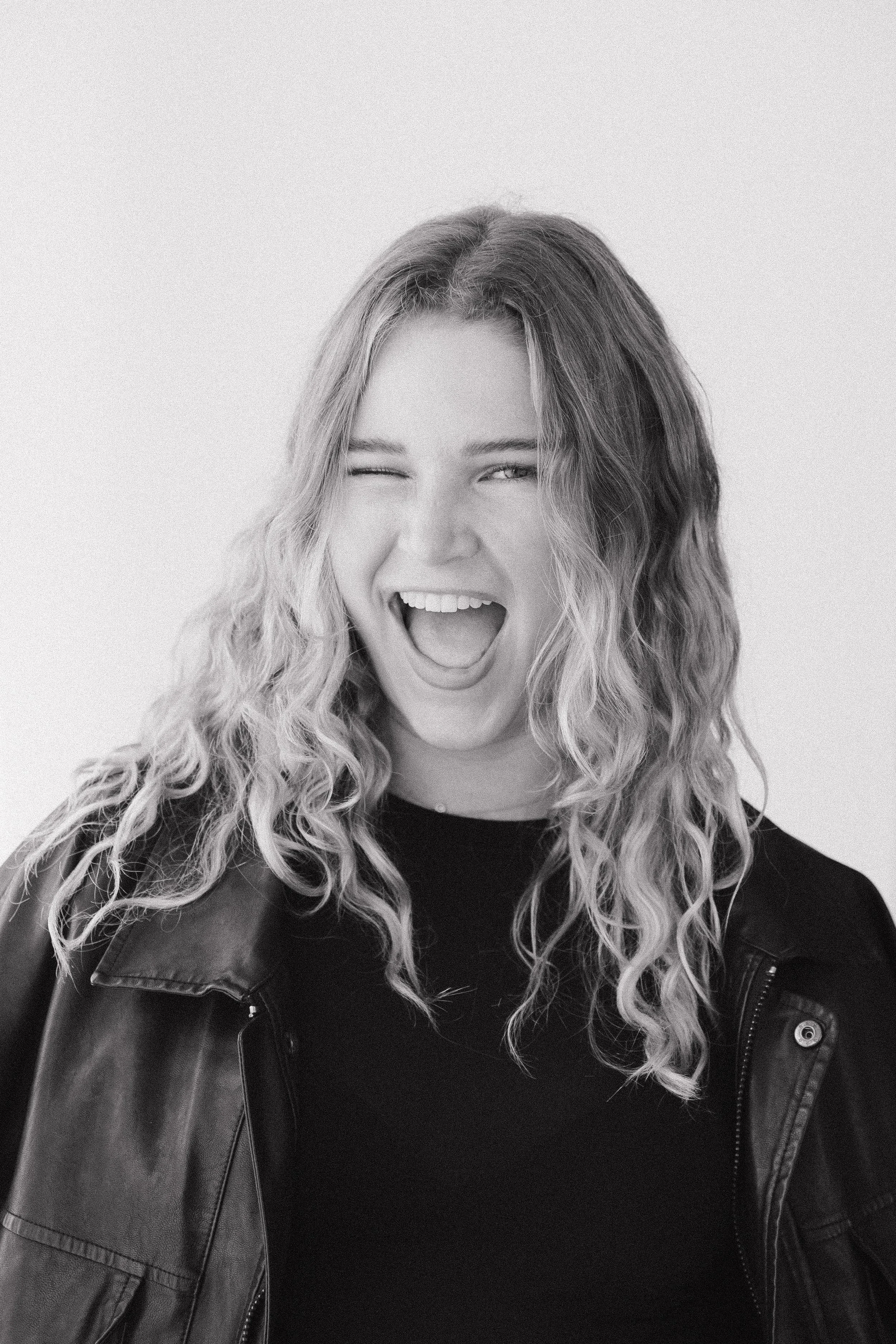 A woman with long curly hair, winking and smiling with her mouth open, wearing a black top and a leather jacket.