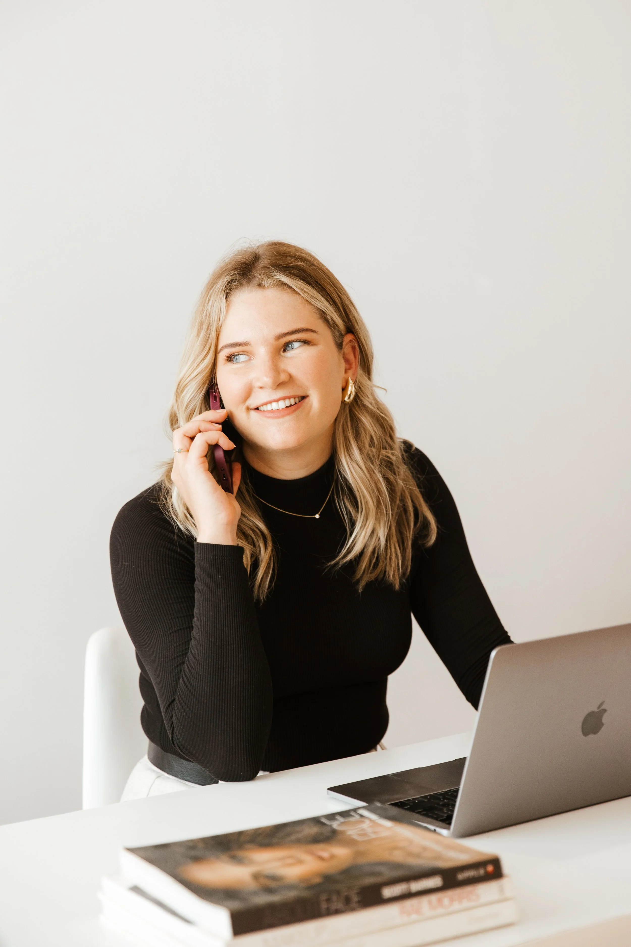 Hannah sitting at a white desk with a silver MacBook, talking on a pink phone, and smiling.