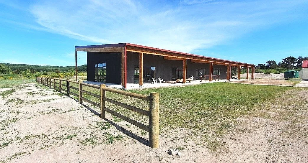 The Wharekai at Tutetawha Marae in Taupo, built by RBS Group. It has a sloped roof, surrounded by a wooden fence, with a mix of grass and dirt in the foreground, and a clear blue sky.