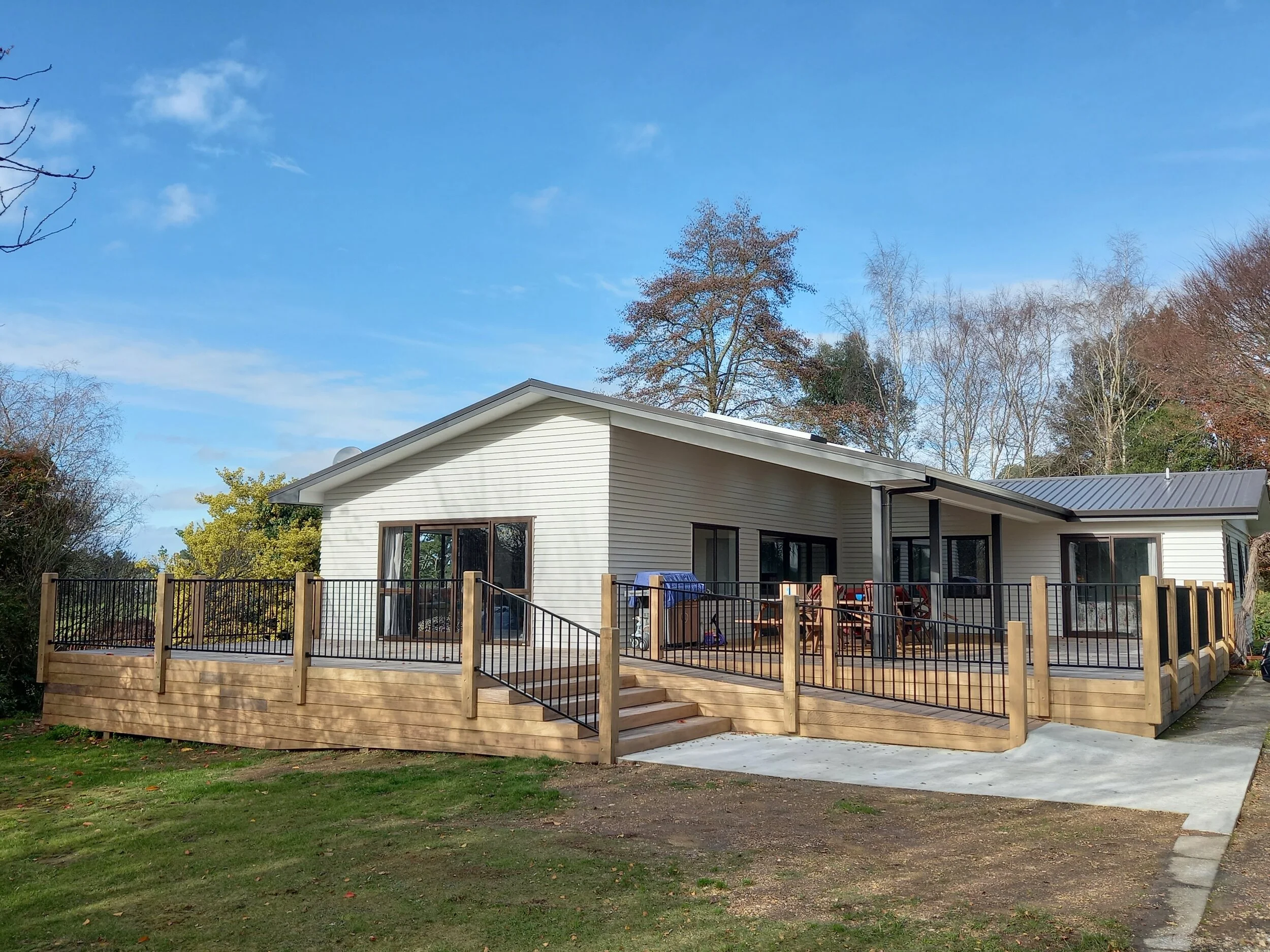 Modern house built by RBS Group in Taupo, with a large wooden deck, black metal railings, sliding glass doors, and a cozy outdoor seating area. Tree-lined background and a partly cloudy sky.