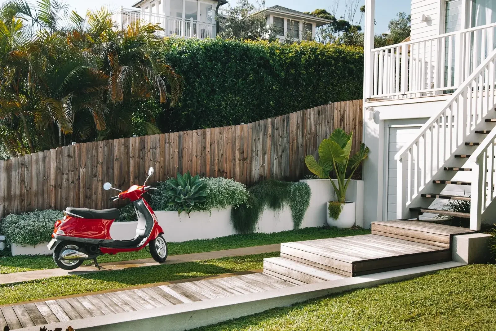 Red scooter parked beside a white house with wooden steps, garden plants, and a wooden fence.