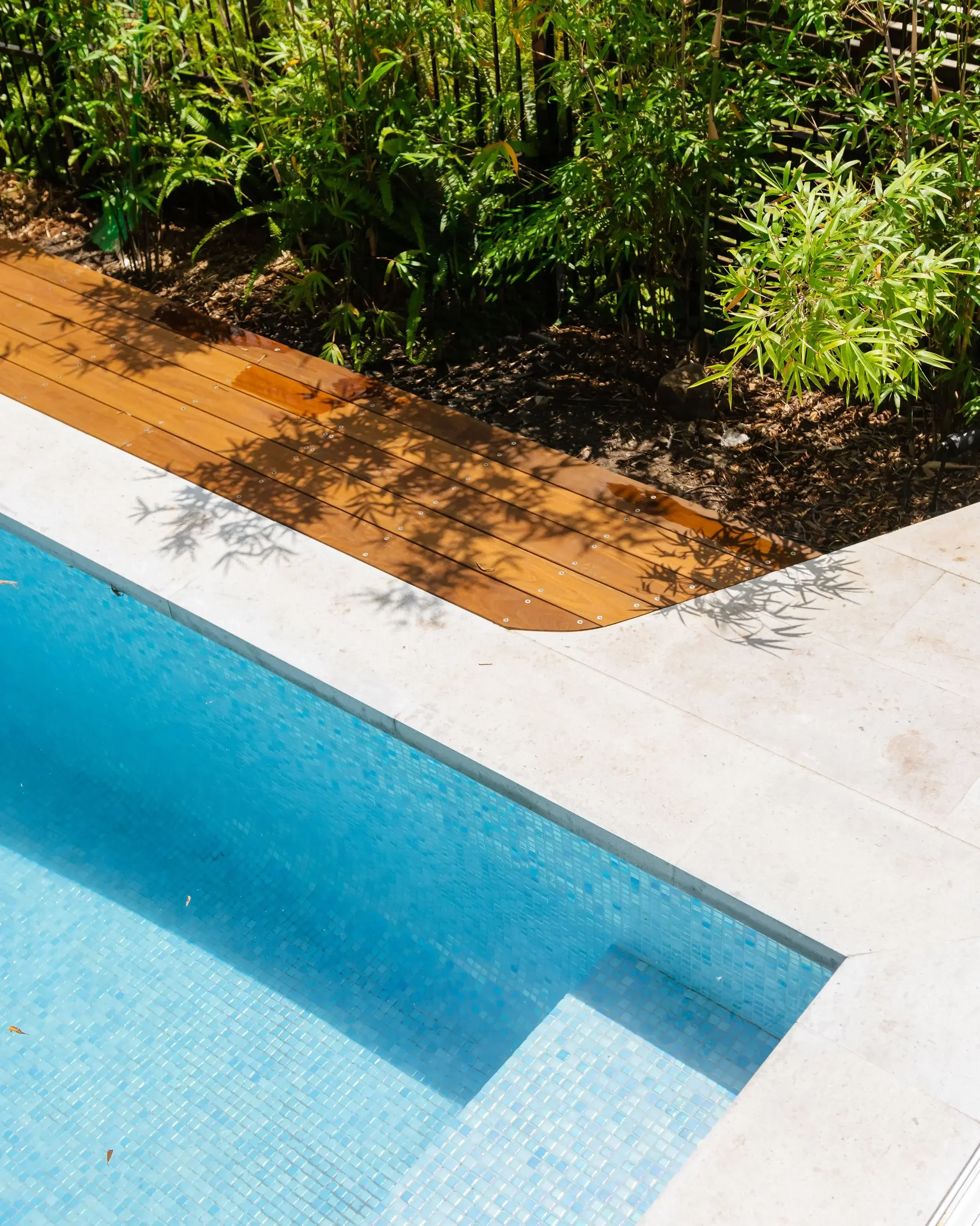Overhead view of a blue‑tiled pool corner with light stone decking and a wooden deck area
