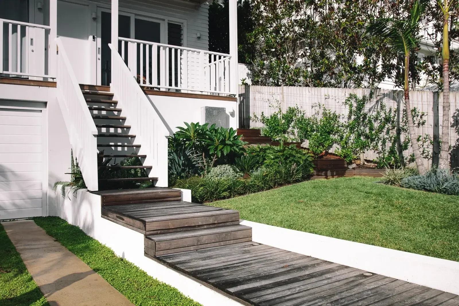 Front yard with wooden steps and a white porch leading up to a house, bordered by green lawn and plants.