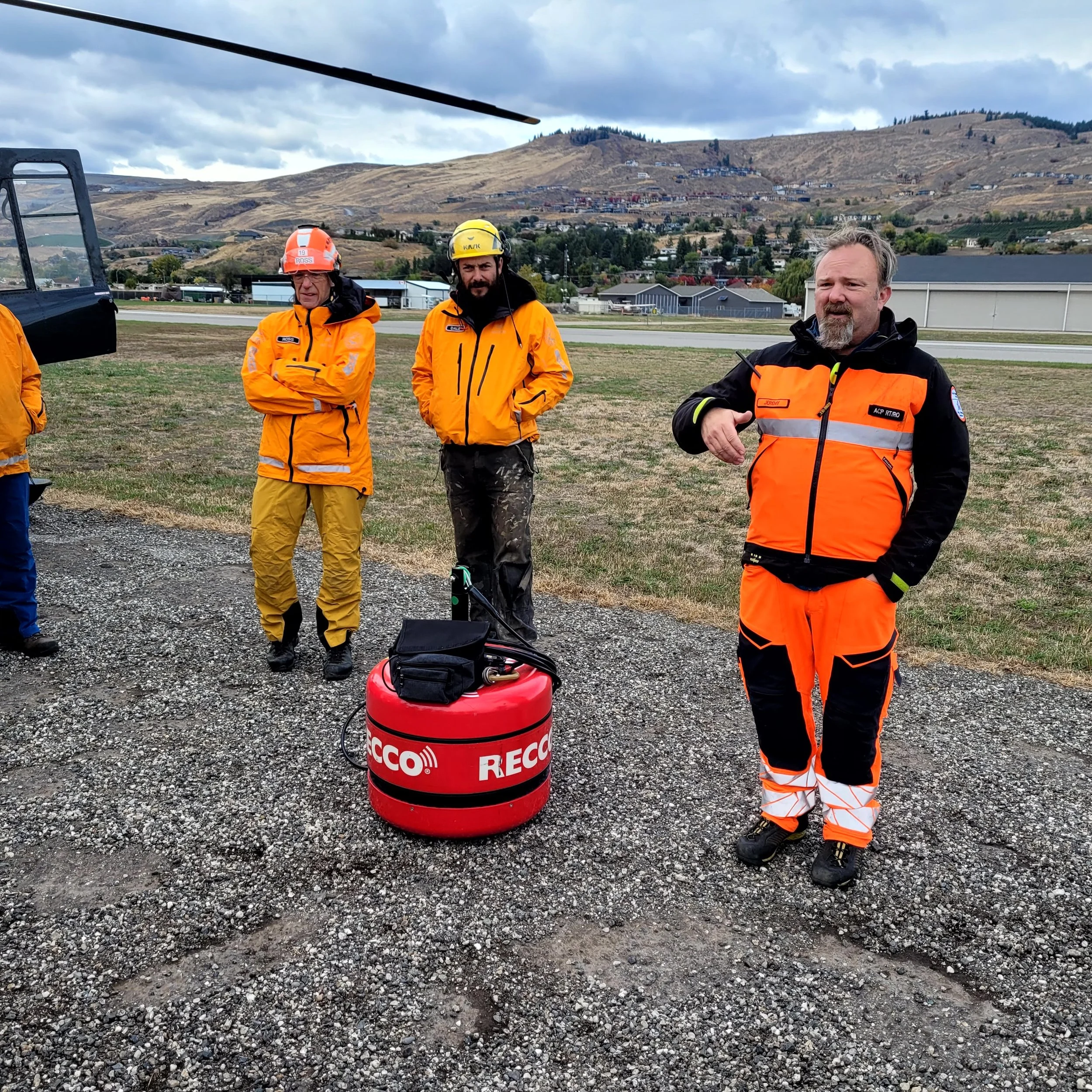 Three emergency responders standing outdoors on gravel, with a landscape of rolling hills and a cloudy sky in the background. One man in the foreground is wearing an orange and black uniform, gesturing with his hand, while other two men in orange jackets and helmets stand behind him. There is equipment on the ground, including a red RECCO device