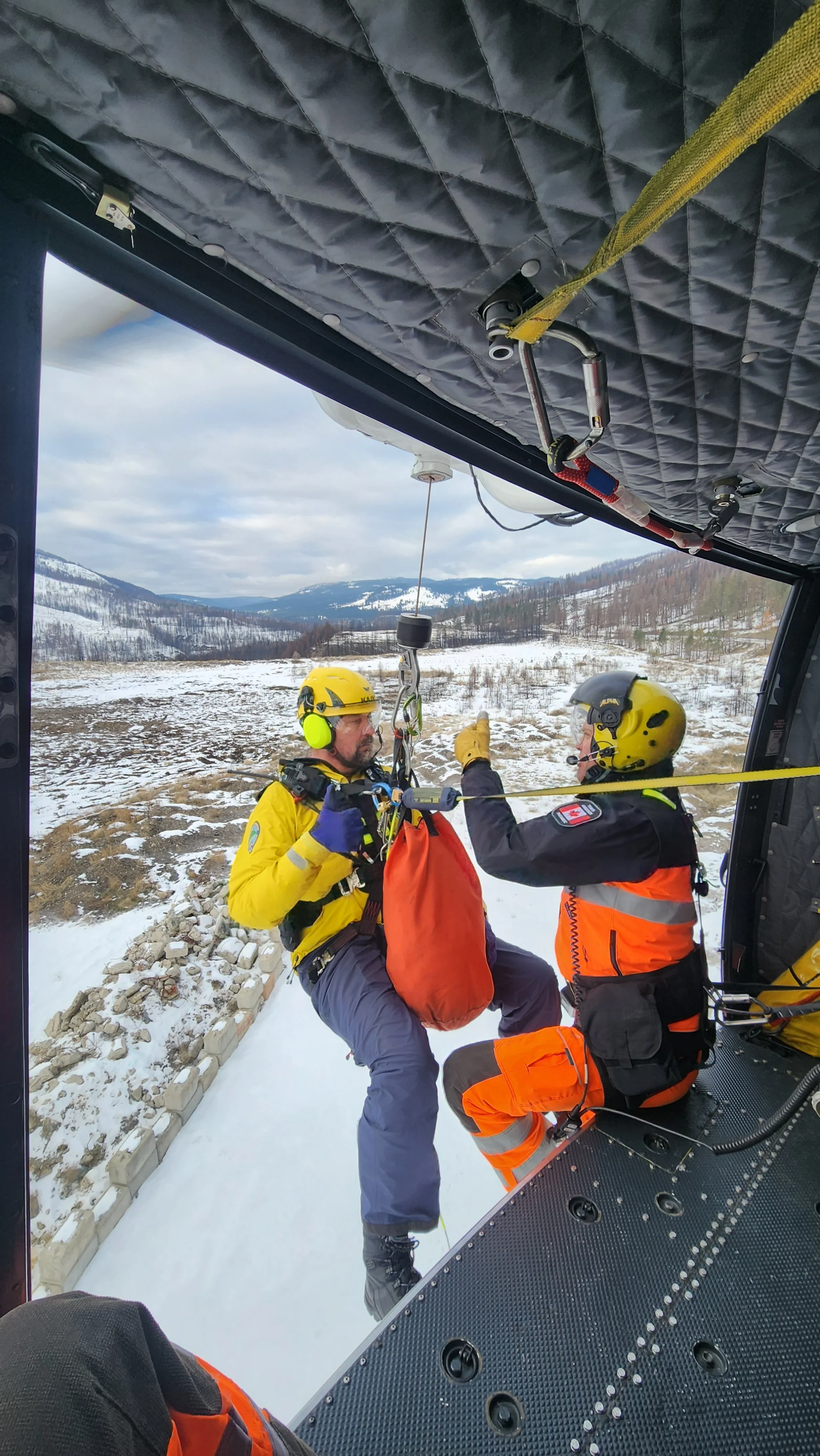 Two rescue workers wearing yellow and orange uniforms, helmets, and harnesses inside a helicopter assisting a person in a snowy outdoor landscape during daytime. Search and Rescue winch training.  Hoist helicopter training.