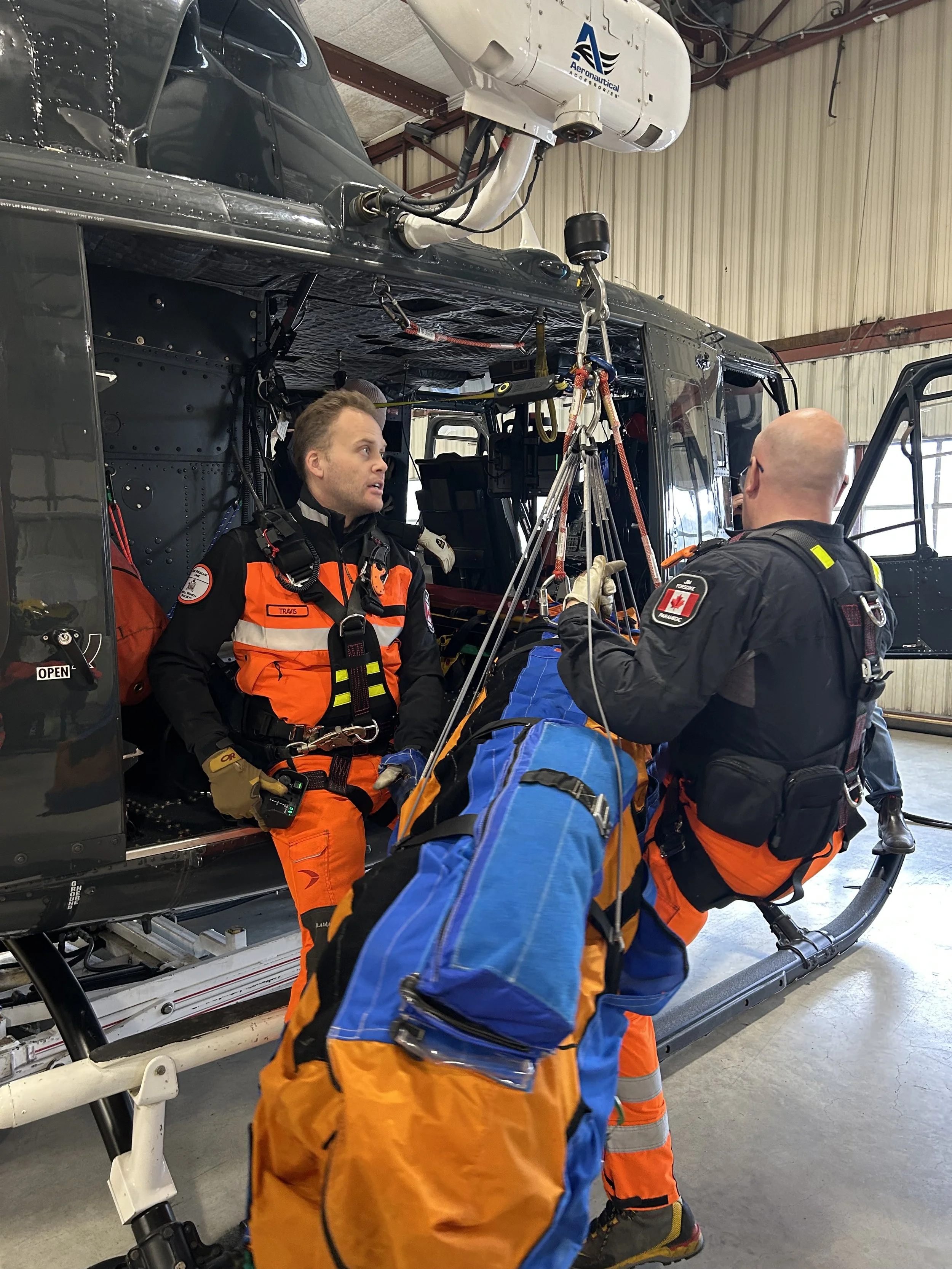Two rescue personnel in orange and black uniforms prepare a patient on a stretcher to load into a helicopter inside a hangar.