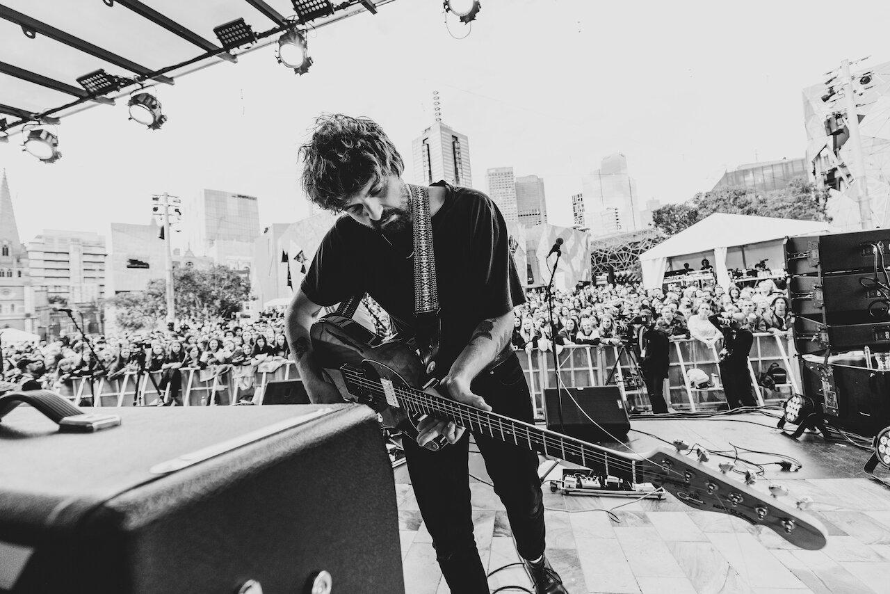 A male guitarist tuning his guitar on an outdoor stage during a concert, with a crowd of people watching and city buildings in the background.