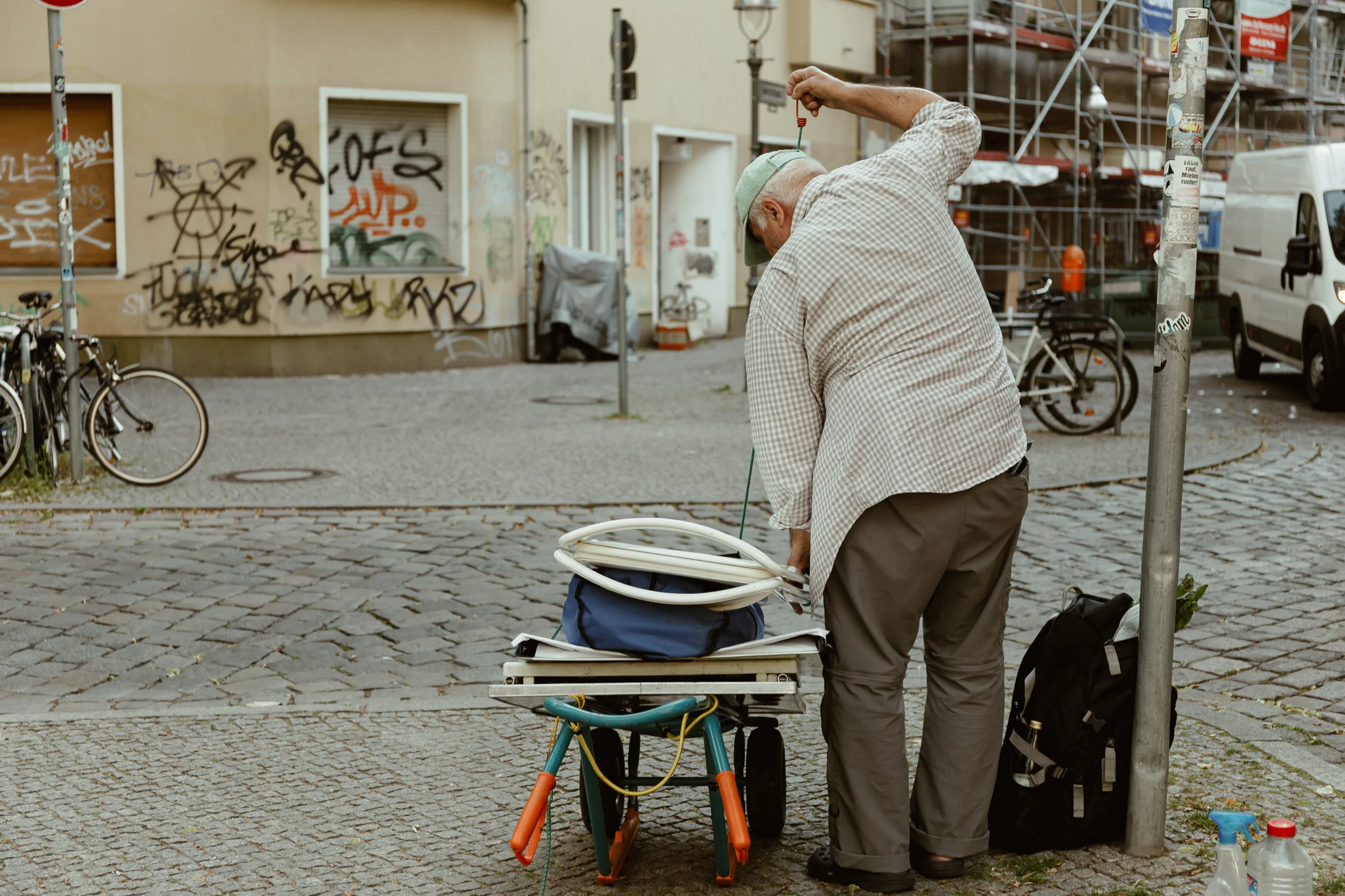 An elderly man with a cap and checkered shirt looks into a cart filled with tubing on a cobblestone street, with graffiti and bicycles in the background.