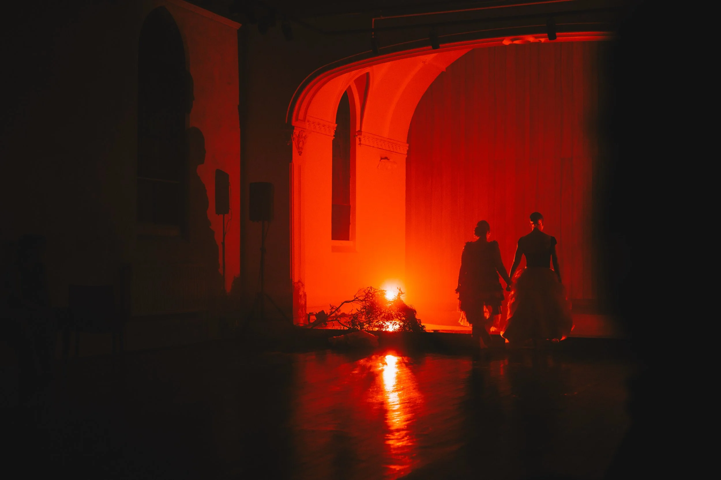 Two people holding hands walking on a stage illuminated with red lighting, with a branch or tree branch on the floor, and shadows of speakers and architecture in the background.