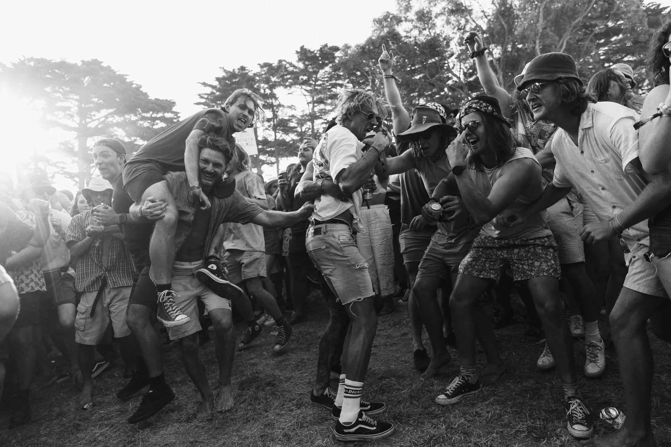 Group of people dancing and celebrating outdoors in a park-like setting with trees in the background, black and white photo.