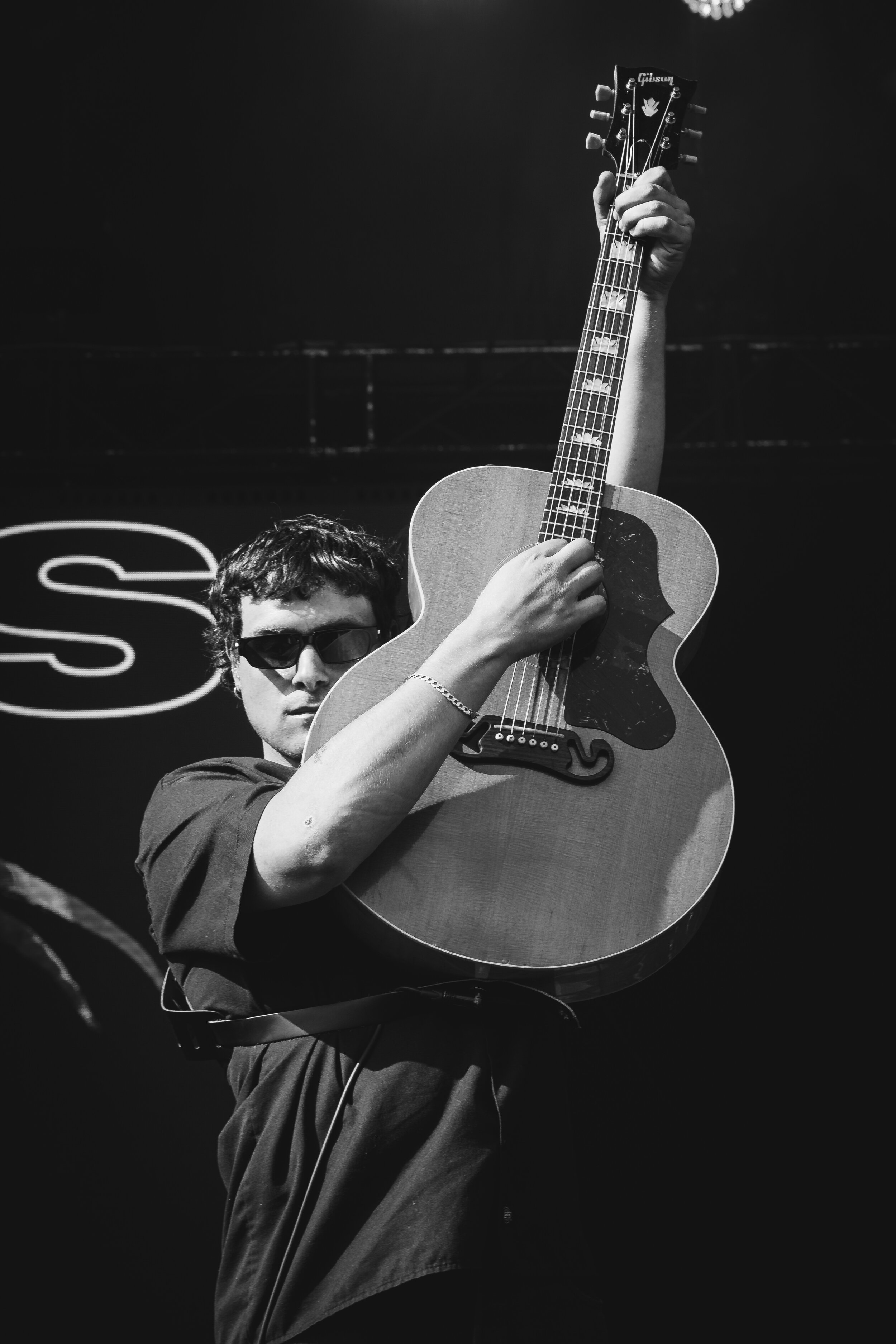 Black and white photo of a person wearing sunglasses playing an acoustic guitar on stage.