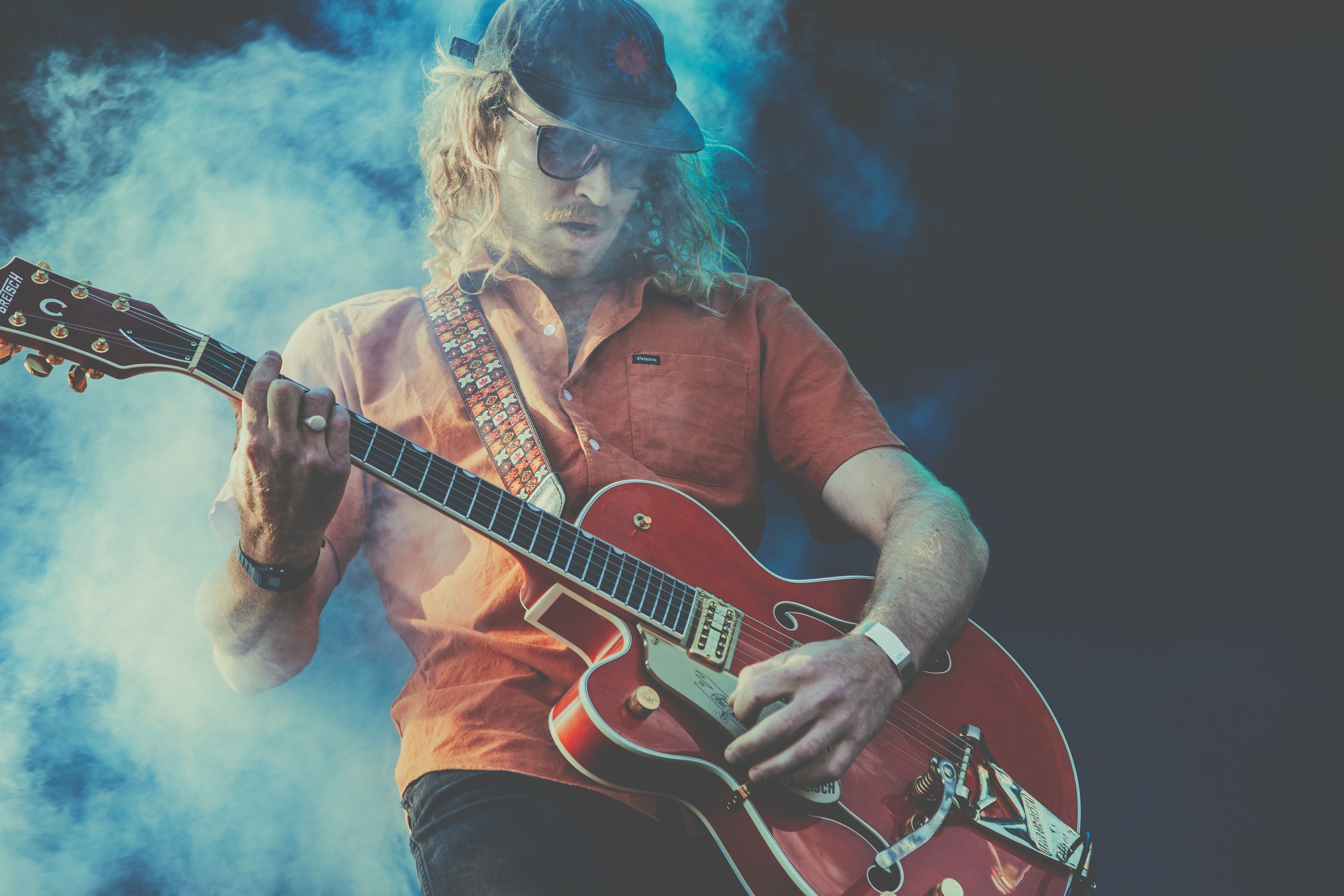A man with curly blonde hair wearing a black cap, sunglasses, an orange shirt, and a wristband is playing an electric guitar on stage with blue smoke around him.