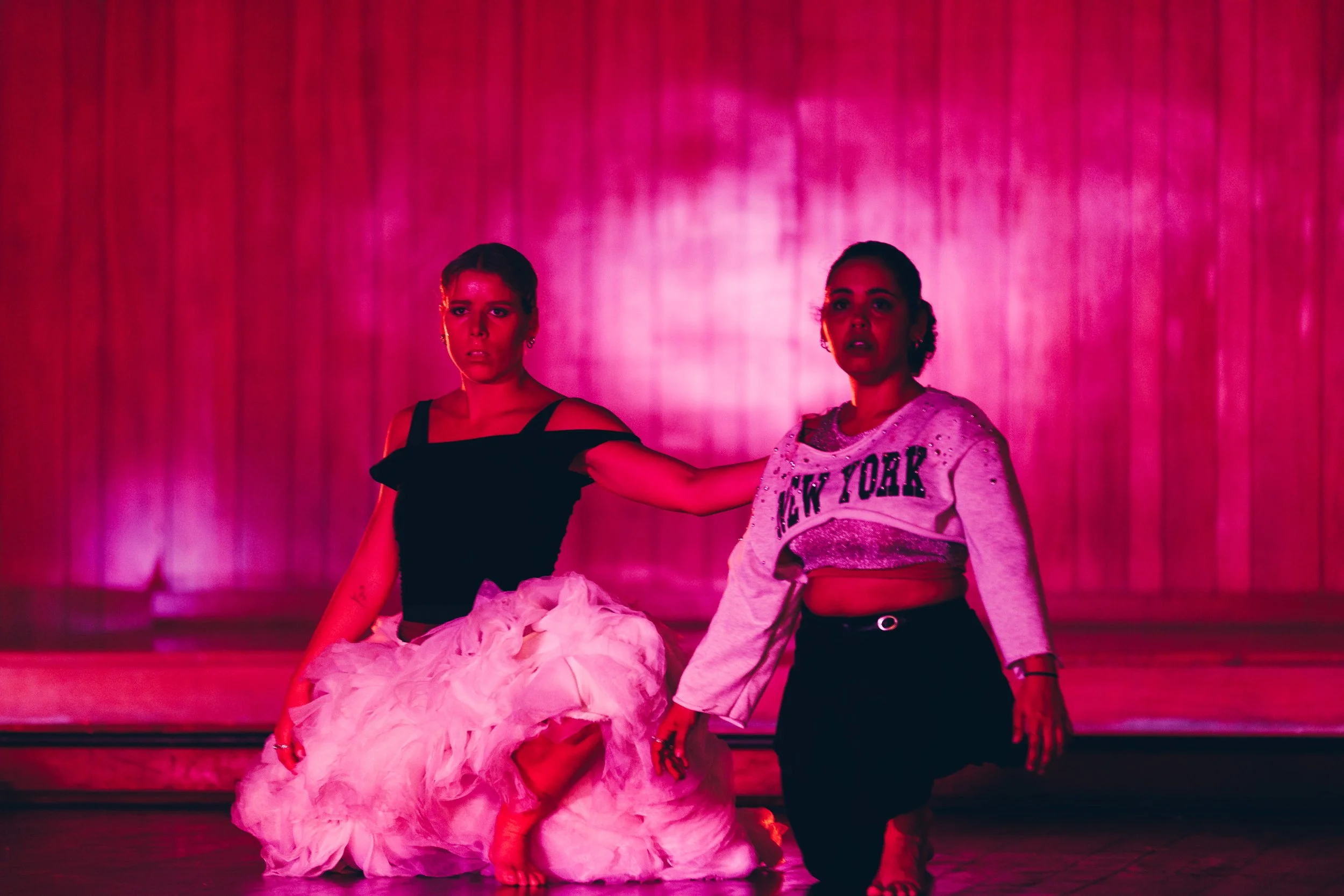 Two women on stage, one sitting in a fluffy pink skirt and black top, the other kneeling in a grey sweatshirt with "New York" written on it, against a pink curtain background.