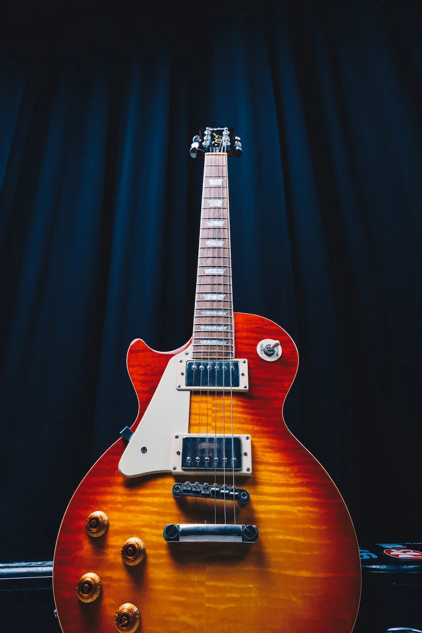 A sunburst finish electric guitar with dual humbucker pickups, four control knobs, and a white pickguard, set against a dark backdrop.