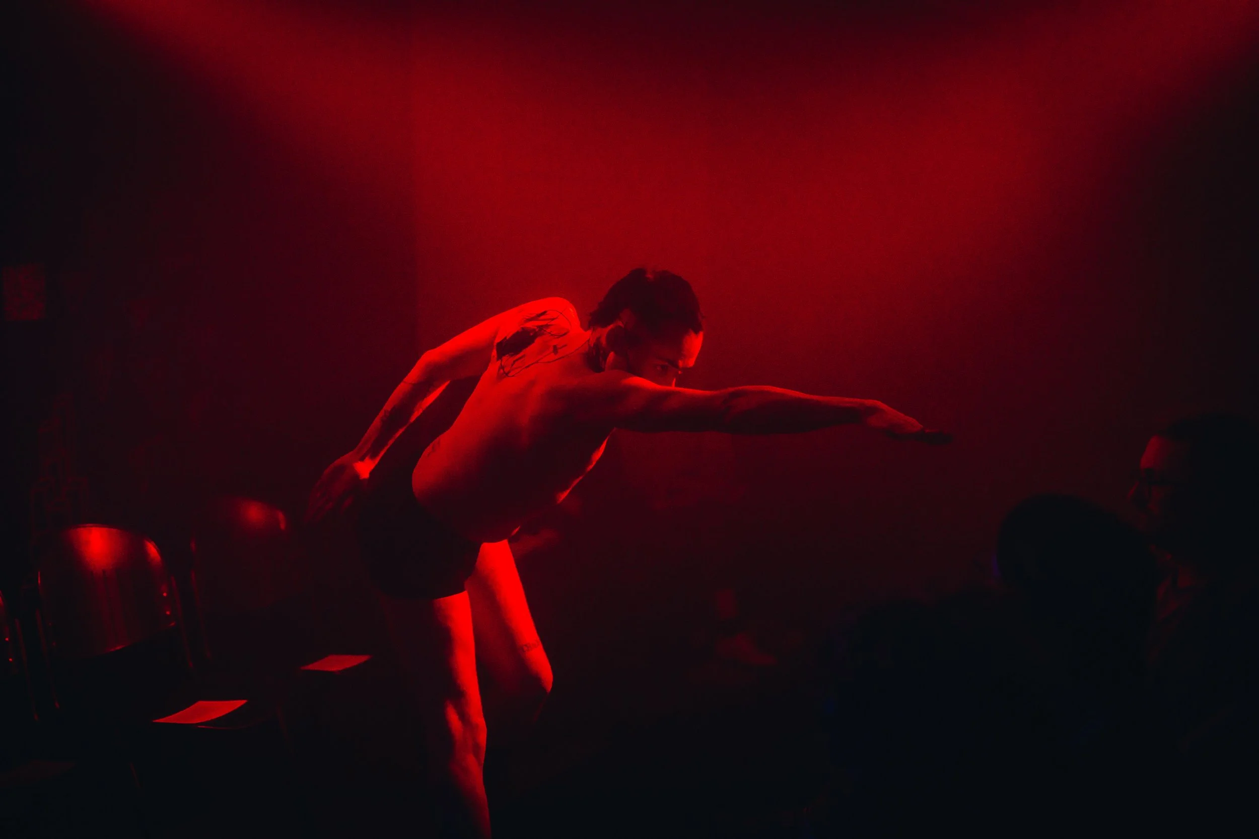 A shirtless male performer in a dark room illuminated by red light, reaching out towards an audience member.
