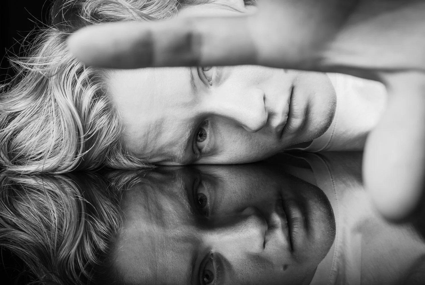 Black and white close-up photograph of a young man lying on a reflective surface with his face and hair reflected. The young man has light-colored, wavy hair and a serious expression, with one eye partially covered by a hand.